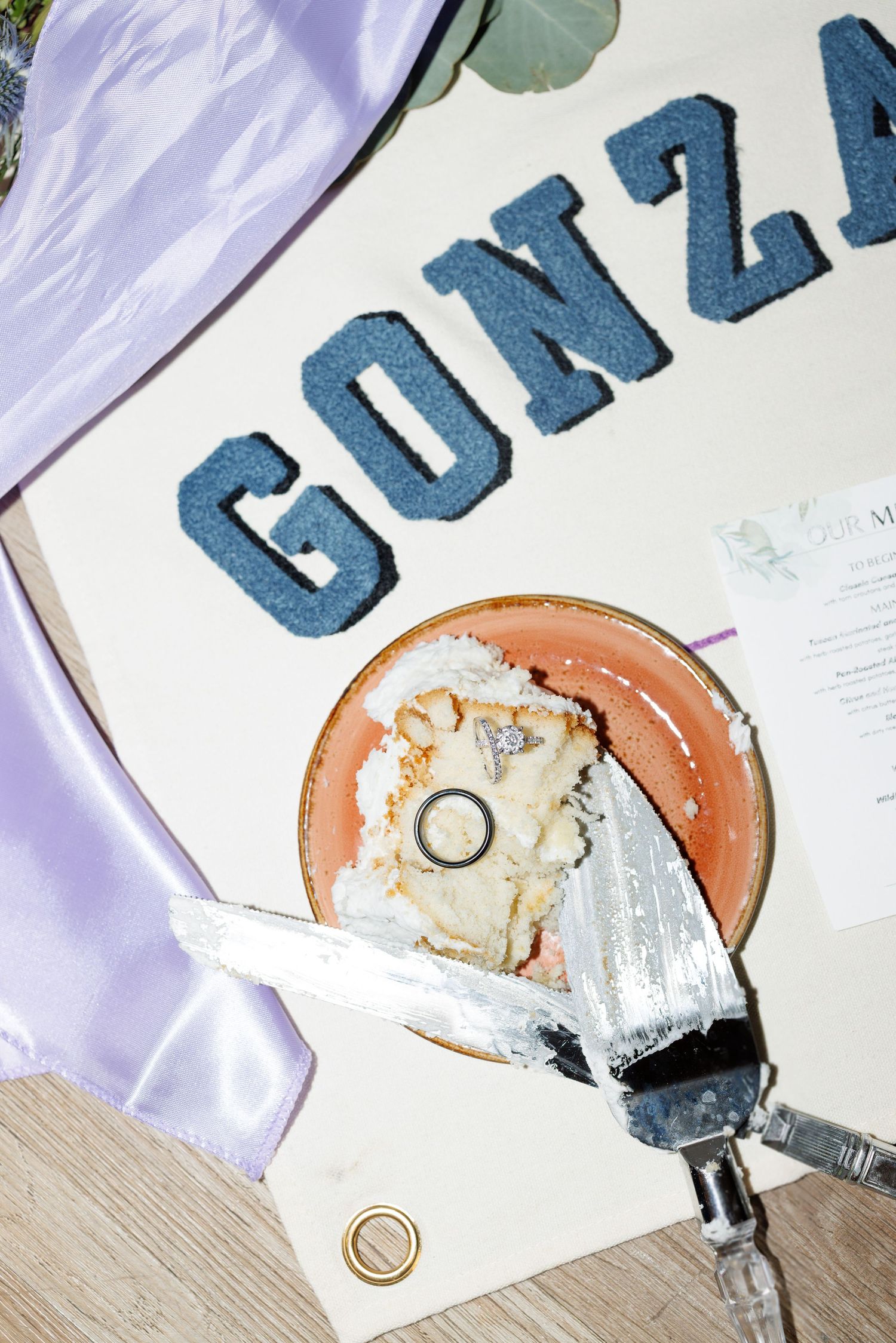Wedding cake slice on plate beside a Gonzaga varsity jacket, cake server, and gold rings on wooden surface.