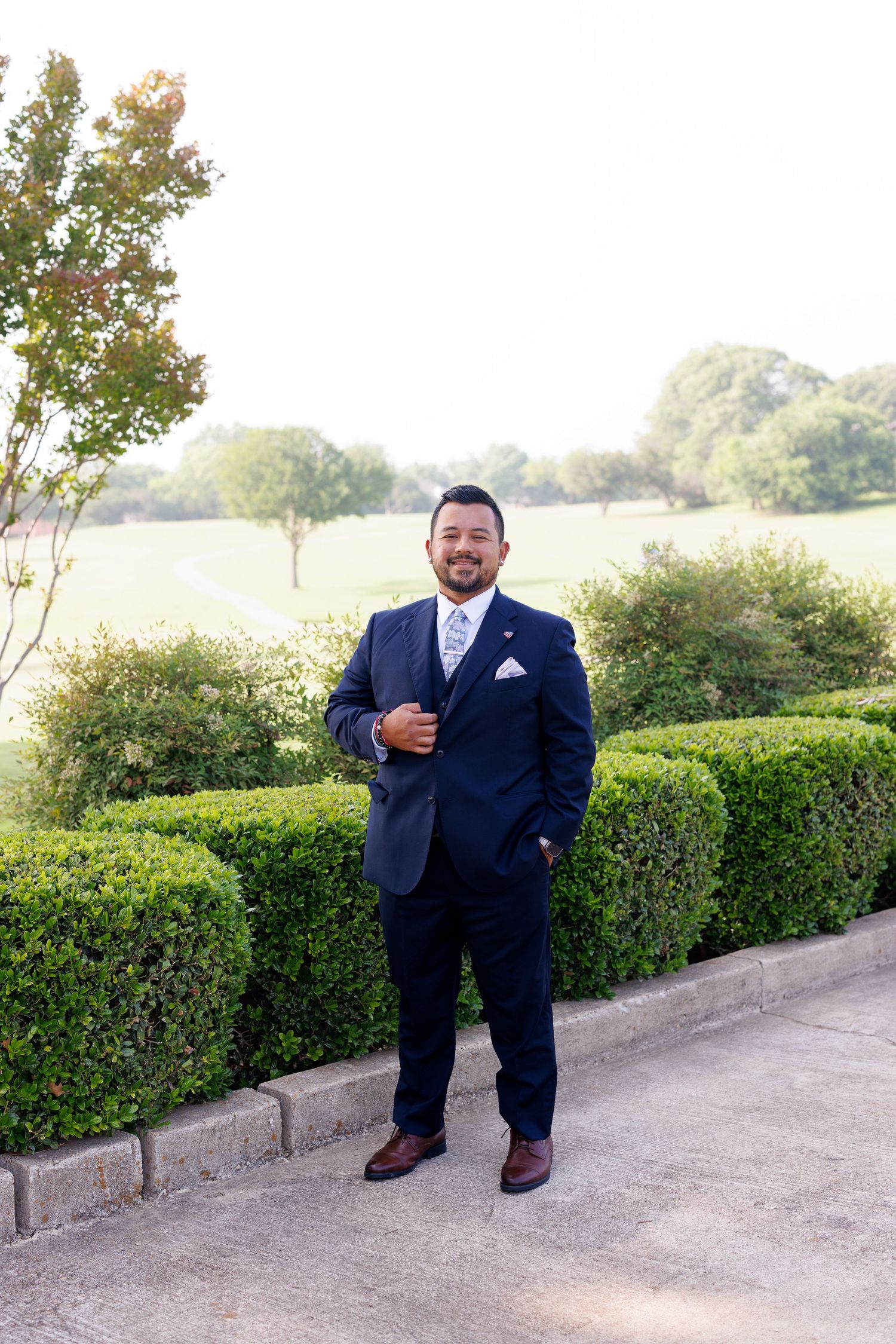 A man in a navy blue suit and tie poses confidently outdoors near manicured hedges and lush green trees.