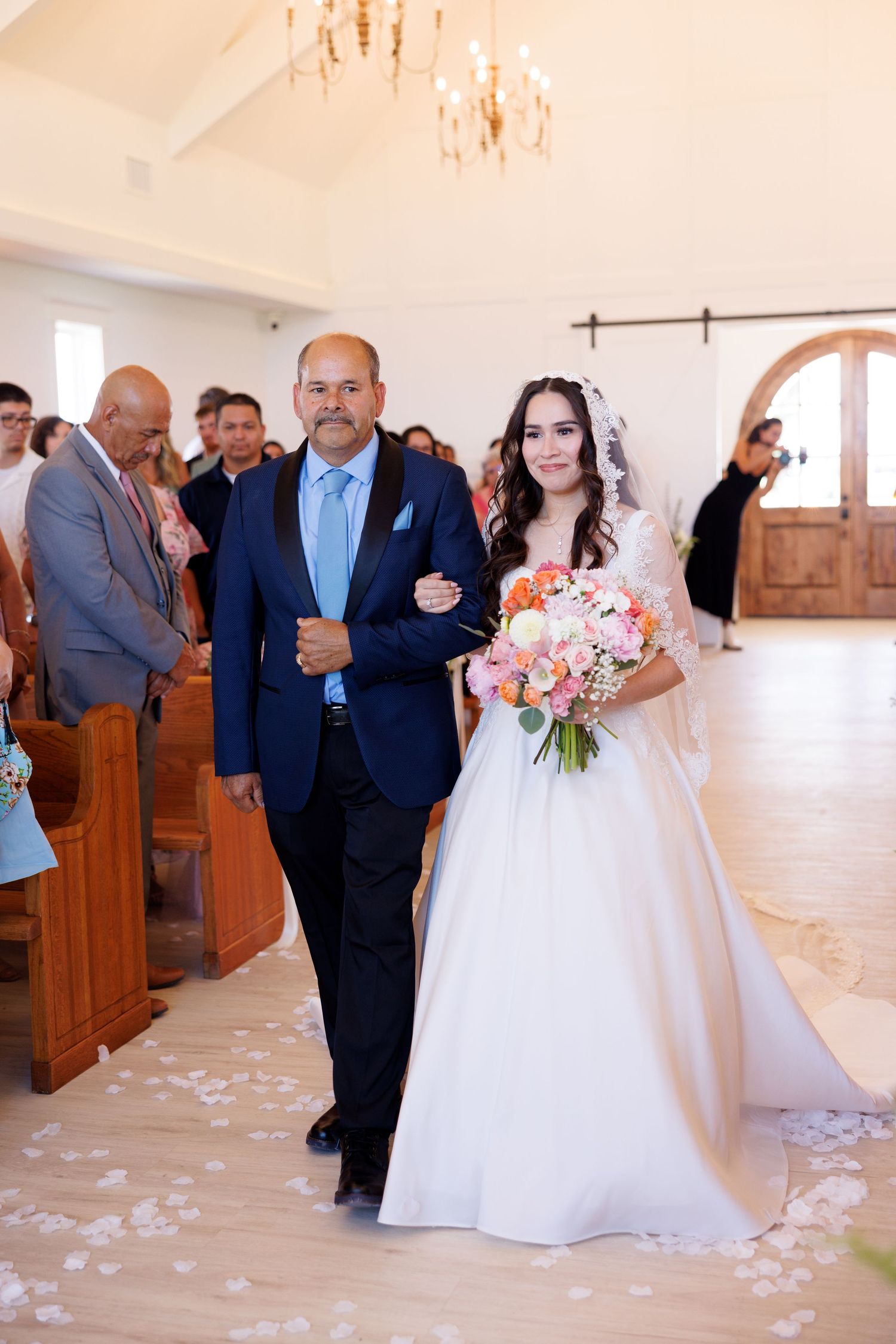 Wedding ceremony procession in an elegant white chapel with scattered rose petals along the aisle.