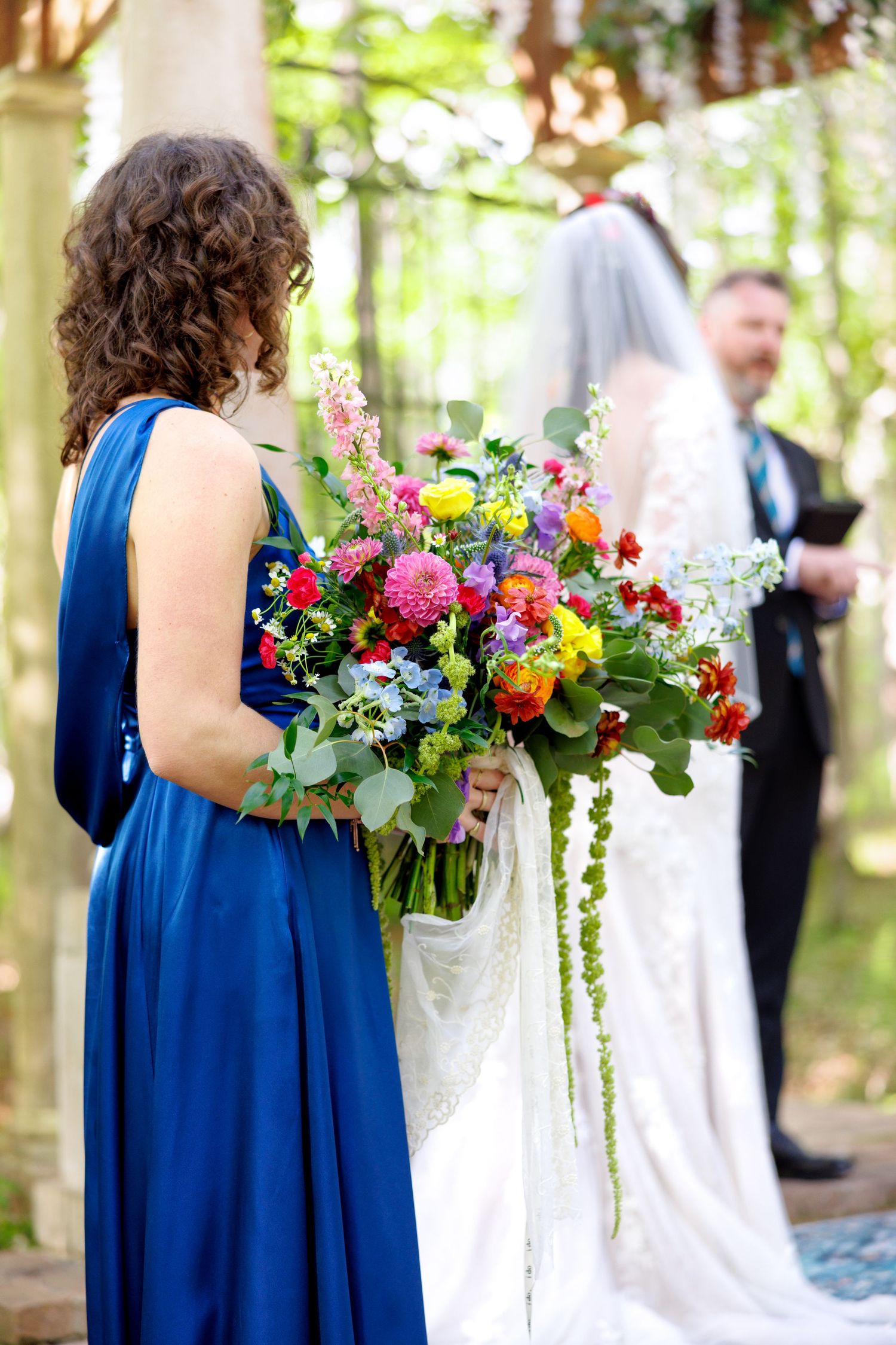 Bridesmaid in blue dress holding vibrant wildflower bouquet during outdoor woodland wedding ceremony.