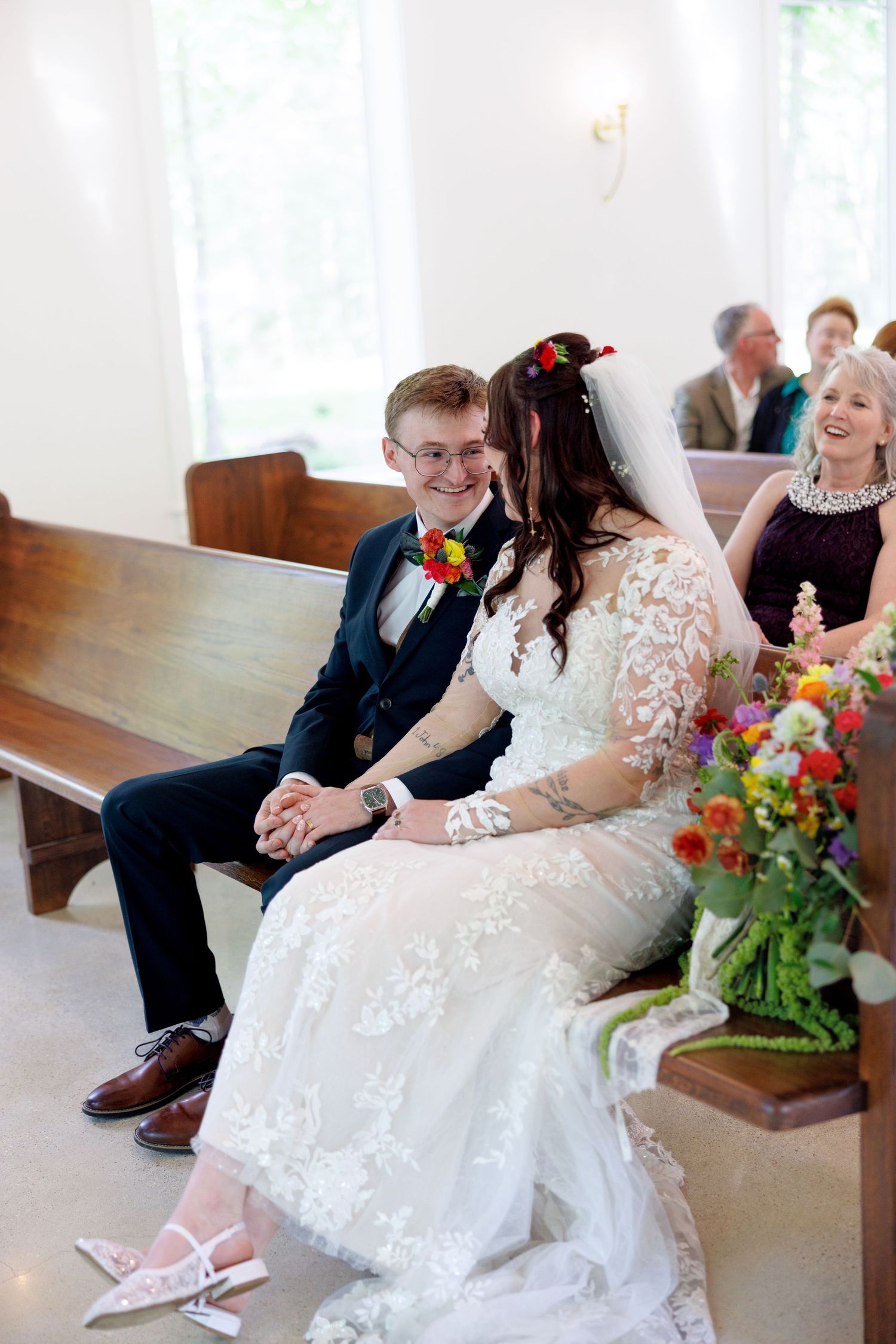 Bride and groom sitting together on church pew, smiling and holding hands during wedding ceremony.