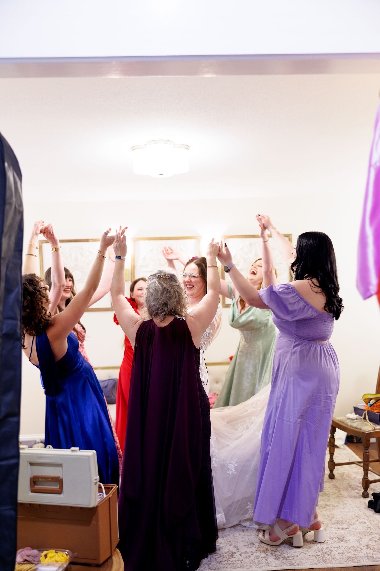 Bridesmaids in colorful gowns celebrate with bride, arms raised joyfully in a warmly lit dressing room.