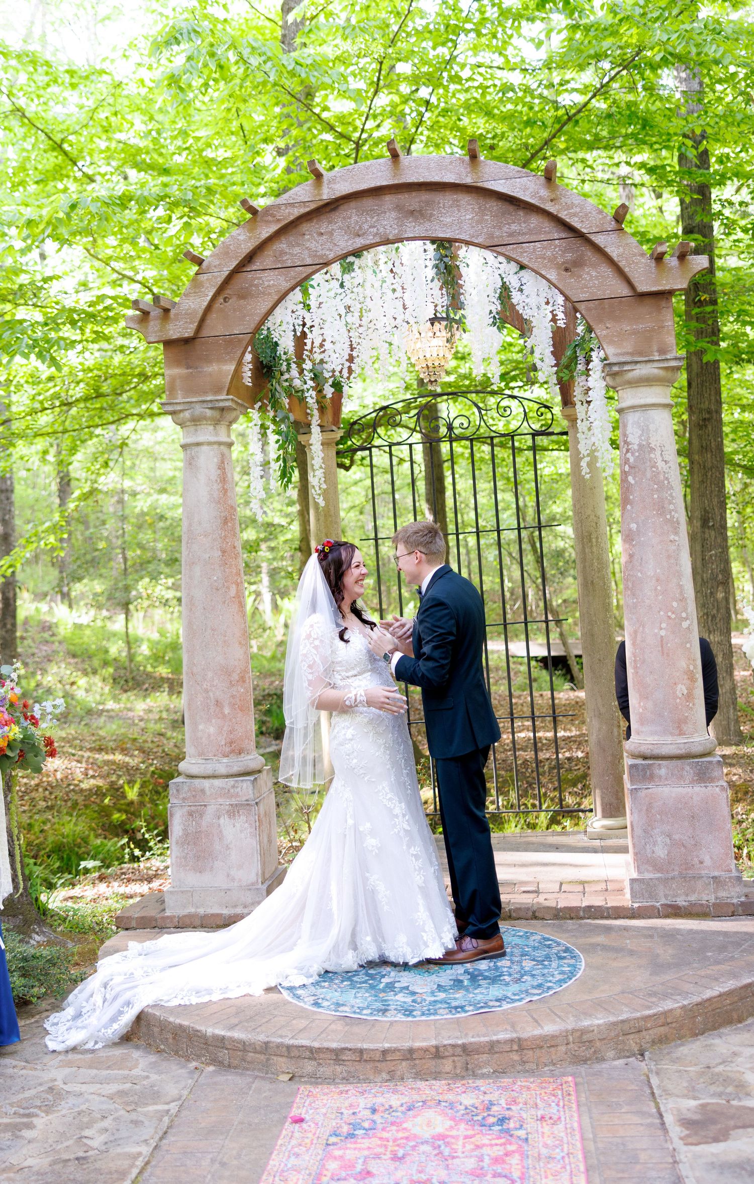 Bride and groom exchange vows under ornate stone arch adorned with white wisteria in forest setting.