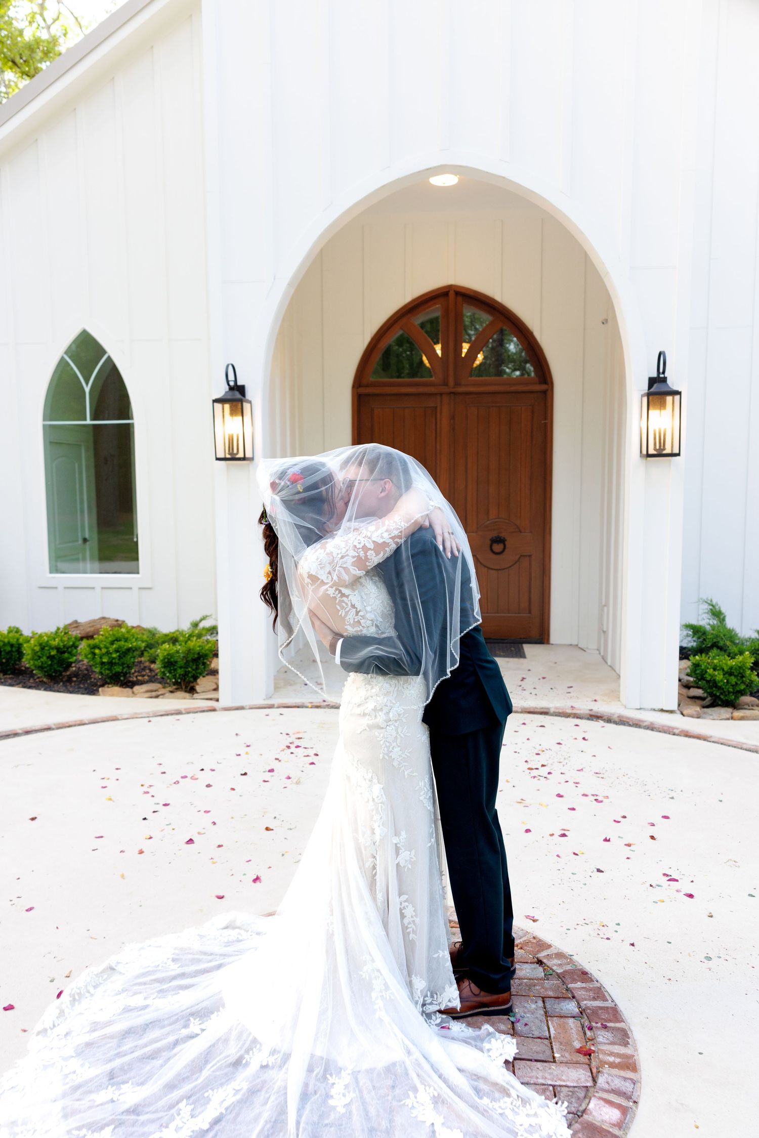Newlyweds share a romantic kiss under the bride's veil outside a white chapel with rose petals on the ground.
