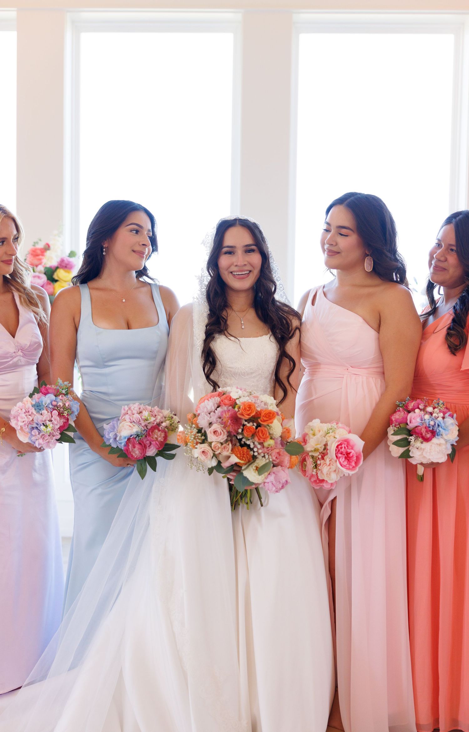 Bridesmaids in pastel dresses hold colorful bouquets while standing in line.