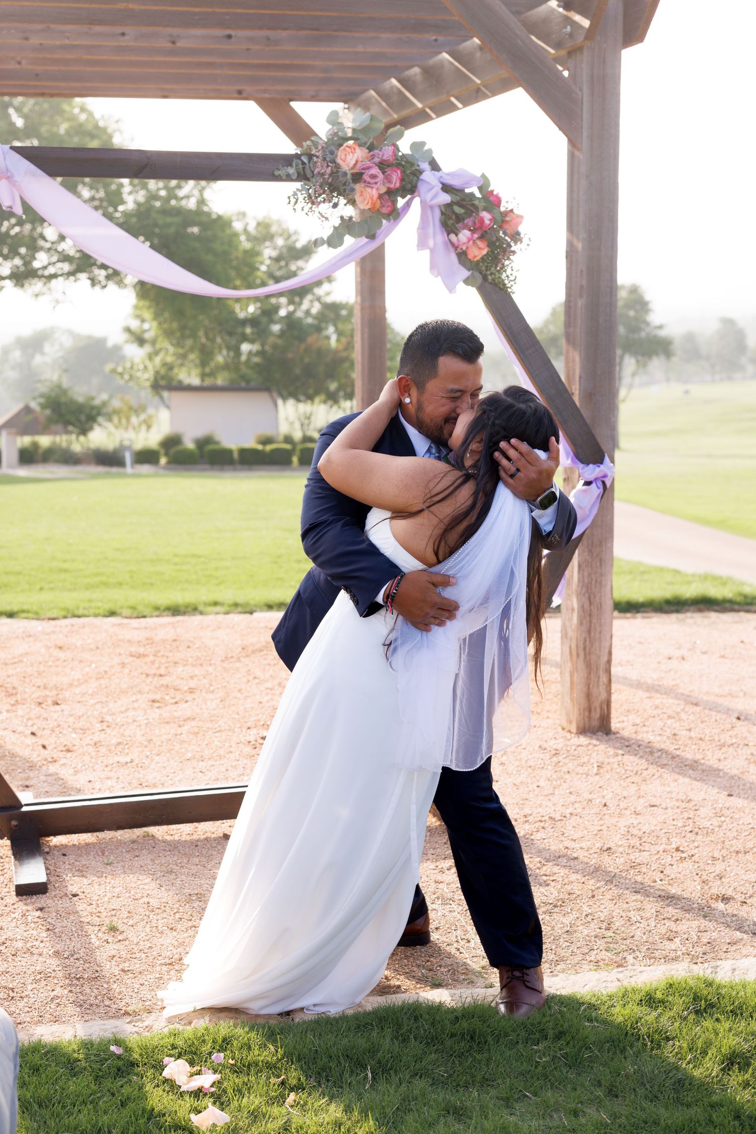 Groom in navy suit dips bride in white gown for first kiss under decorated wooden pergola.