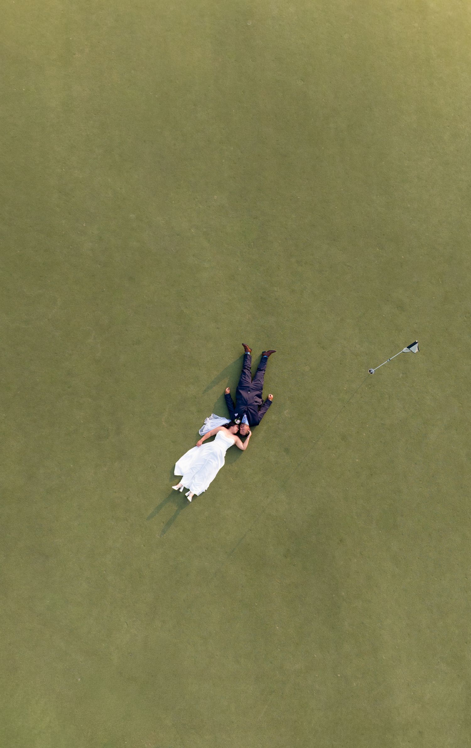 Aerial drone photo of bride and groom lying on green golf course grass in wedding attire, romantic overhead perspective.