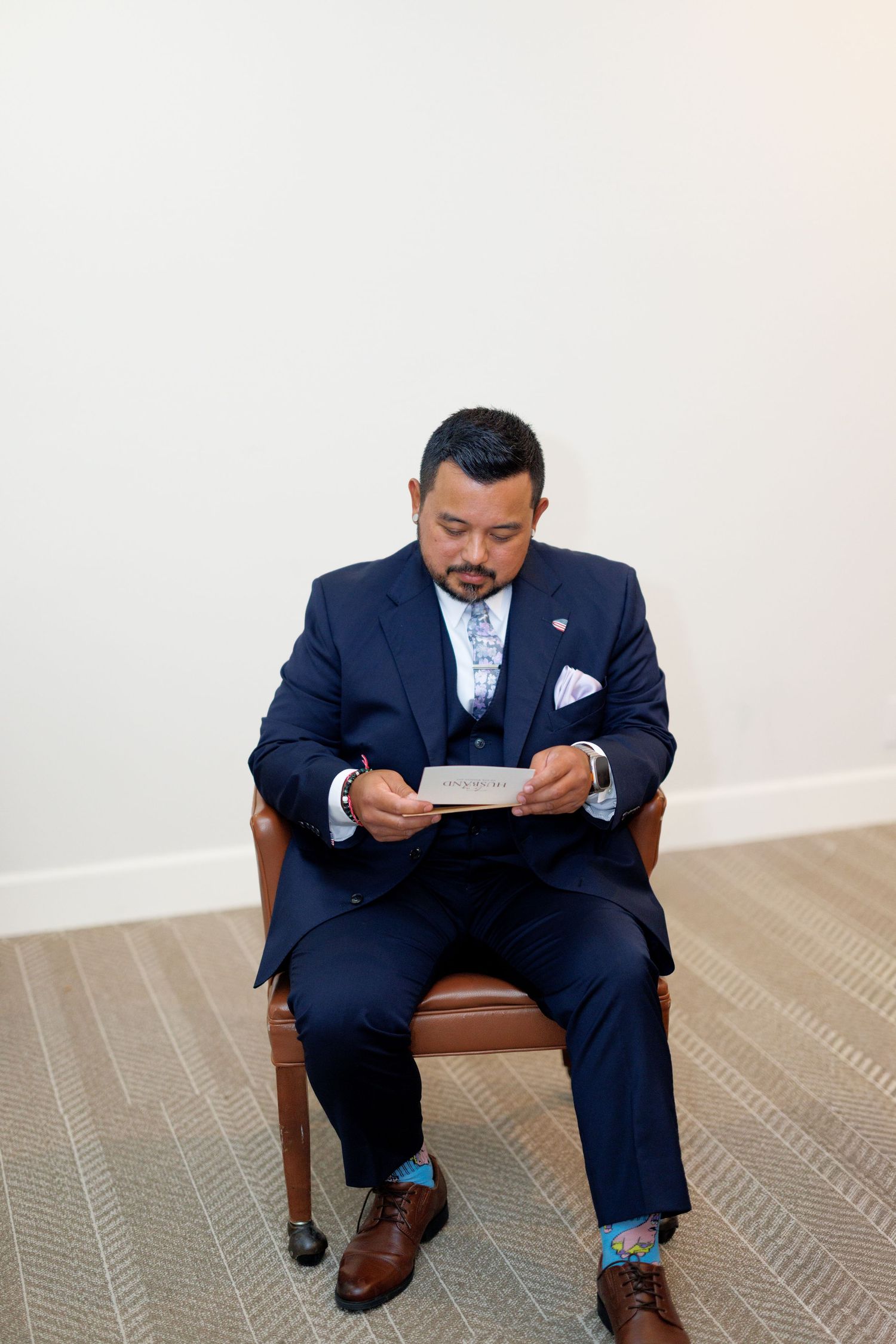 Groom in navy suit reading a card while seated in a leather chair before his wedding ceremony.