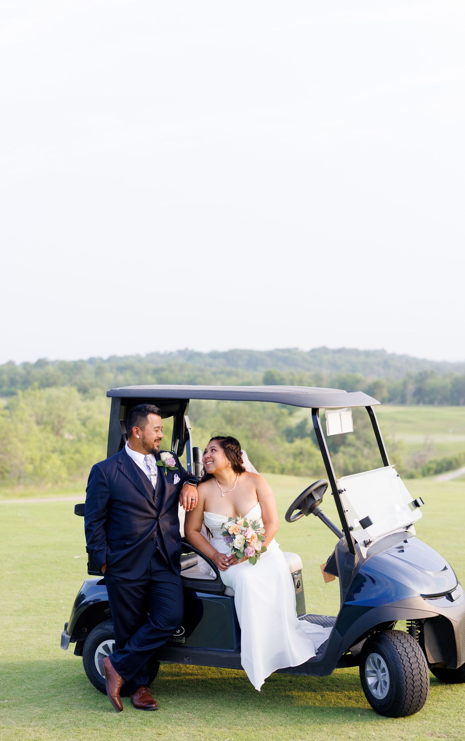 Bride and groom posing by a golf cart on a lush green course, gazing at each other, bride holding a floral bouquet.