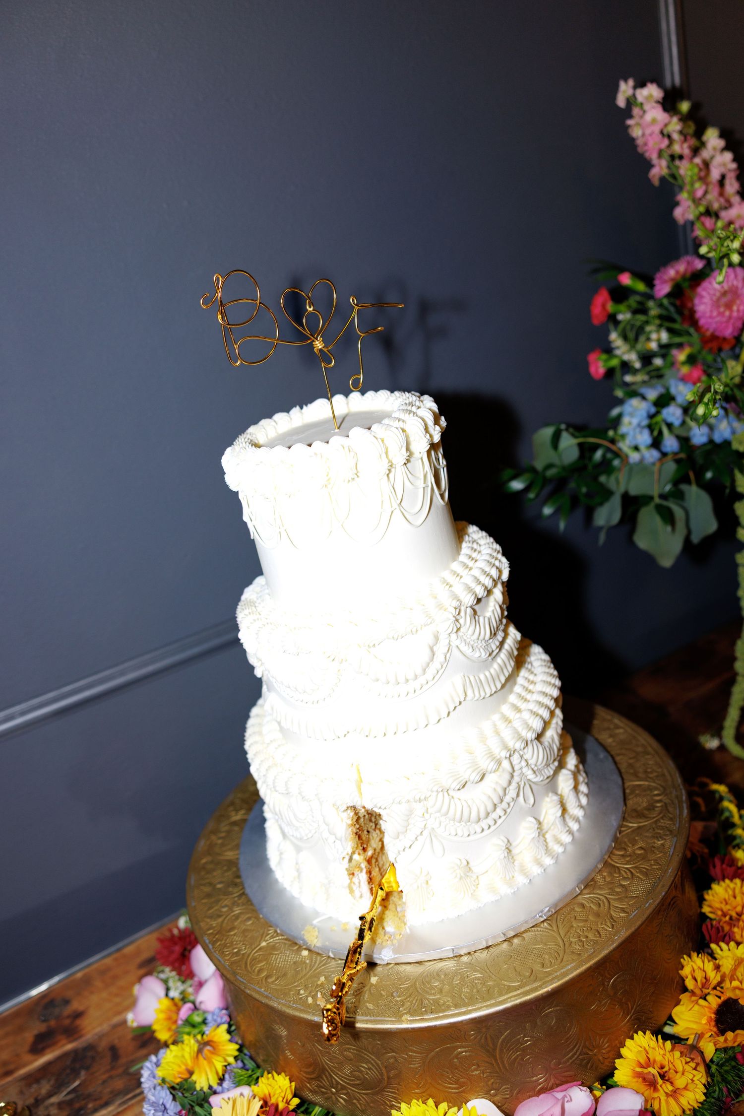 Three-tier white wedding cake with gold wire topper on gold stand, surrounded by colorful flowers against gray wall.