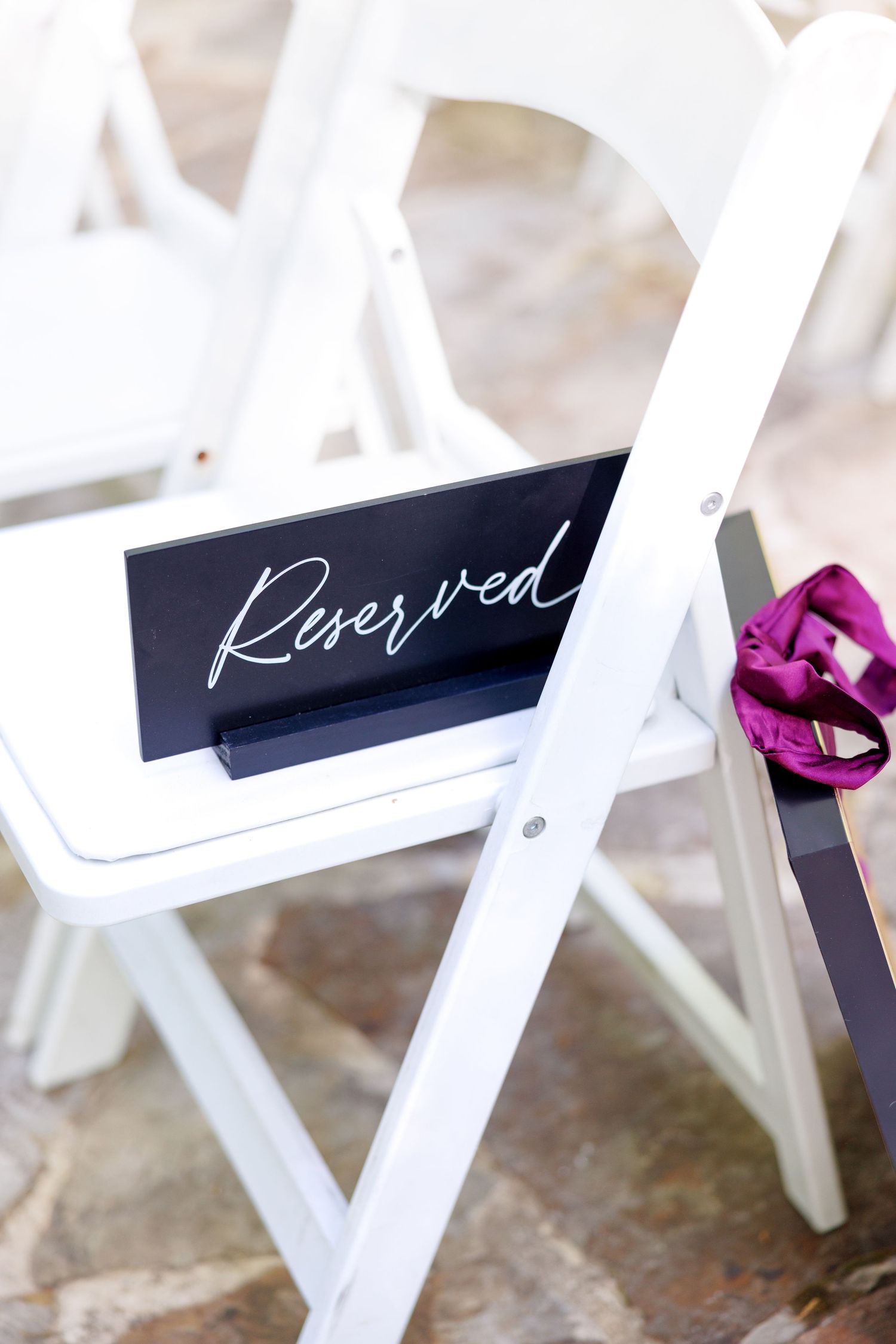 Black reserved sign on white folding chair with purple ribbon bow at outdoor wedding ceremony.