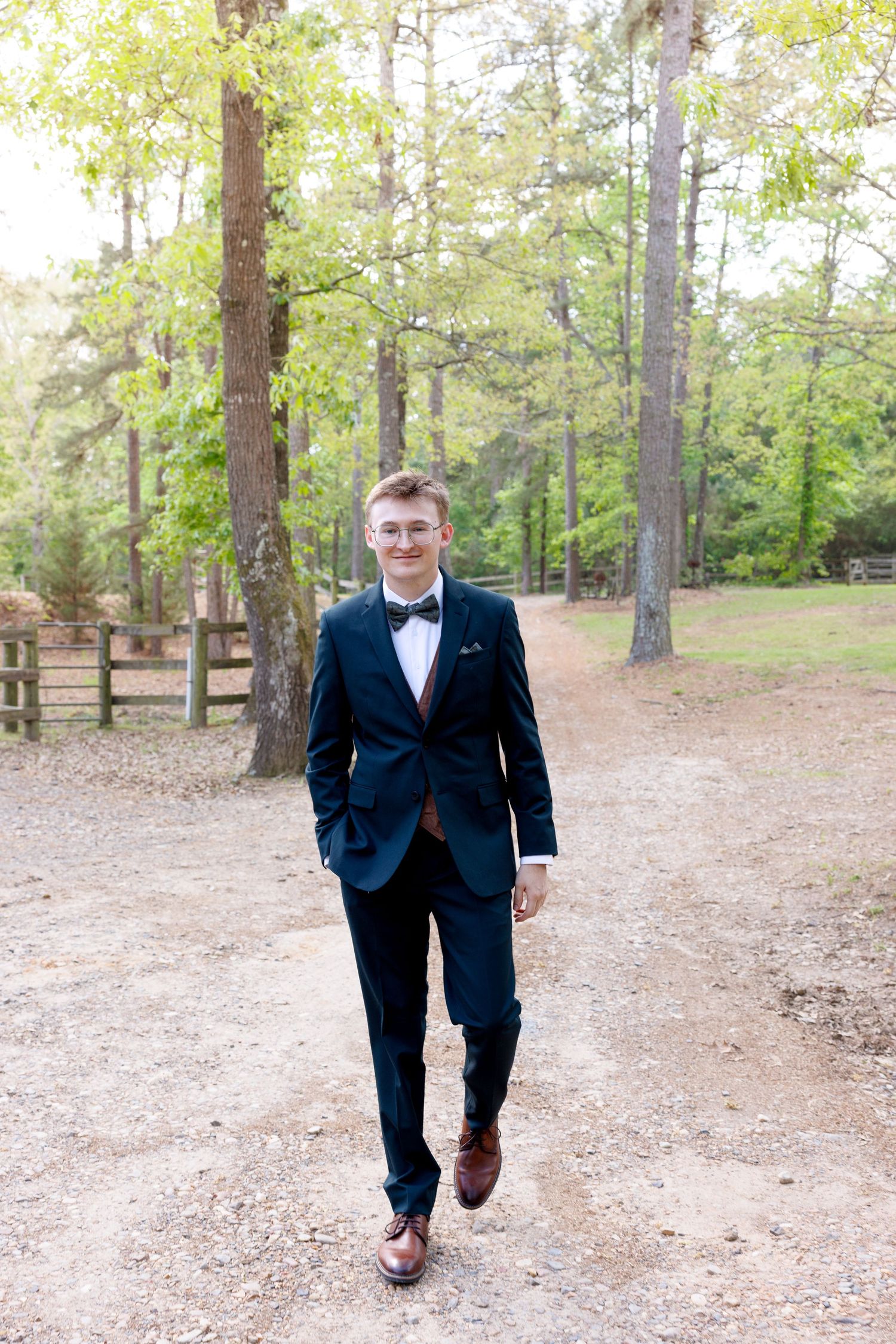 Groom in navy tuxedo with bow tie walks along gravel path surrounded by tall trees.