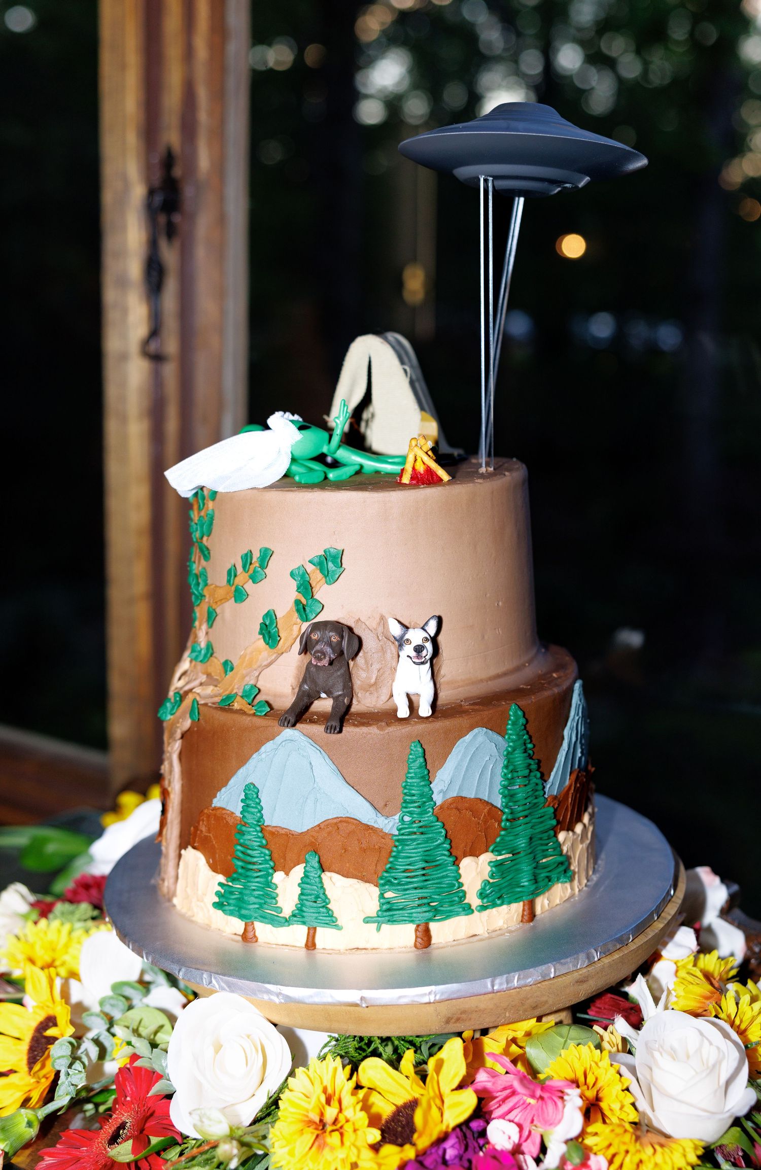 Two-tier camping themed wedding cake with UFO topper, dogs, pine trees, and mountains, displayed on floral arrangement.