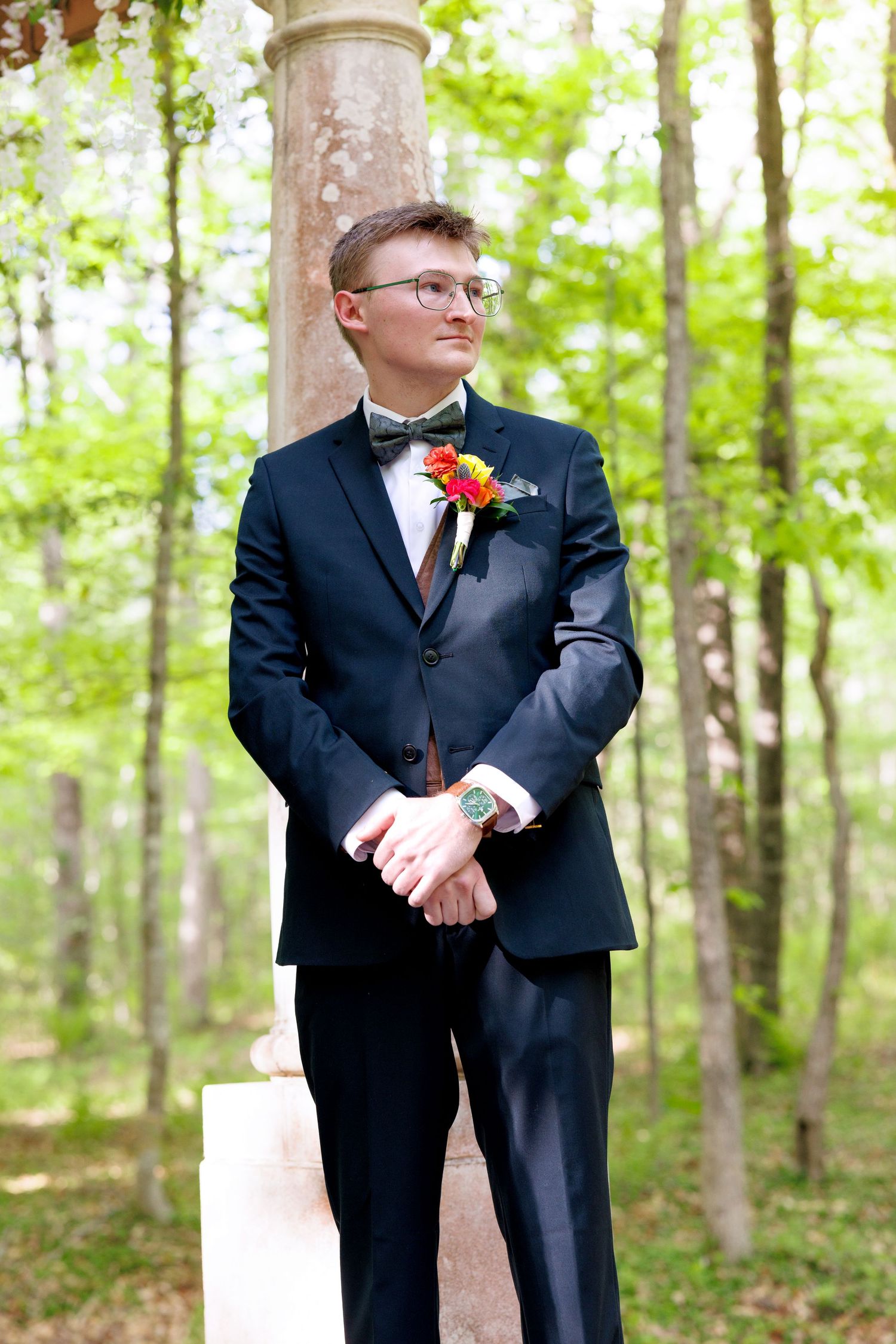 Elegant groom in navy suit with colorful boutonniere and bow tie standing outdoors among trees at wedding.