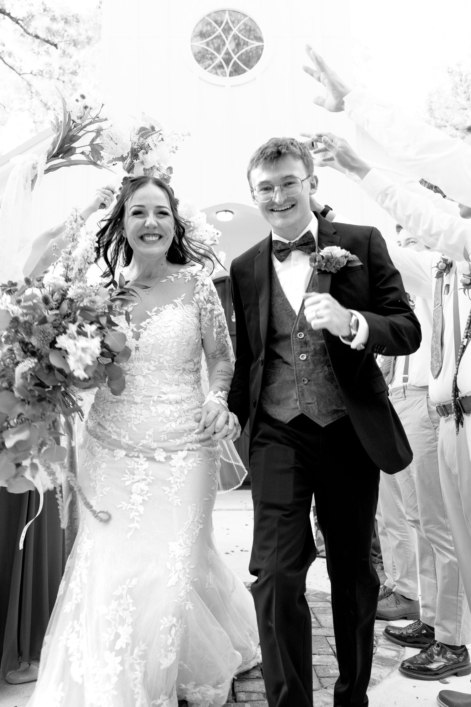 Joyful bride and groom exit church after wedding ceremony, bride holding floral bouquet in elegant lace gown.