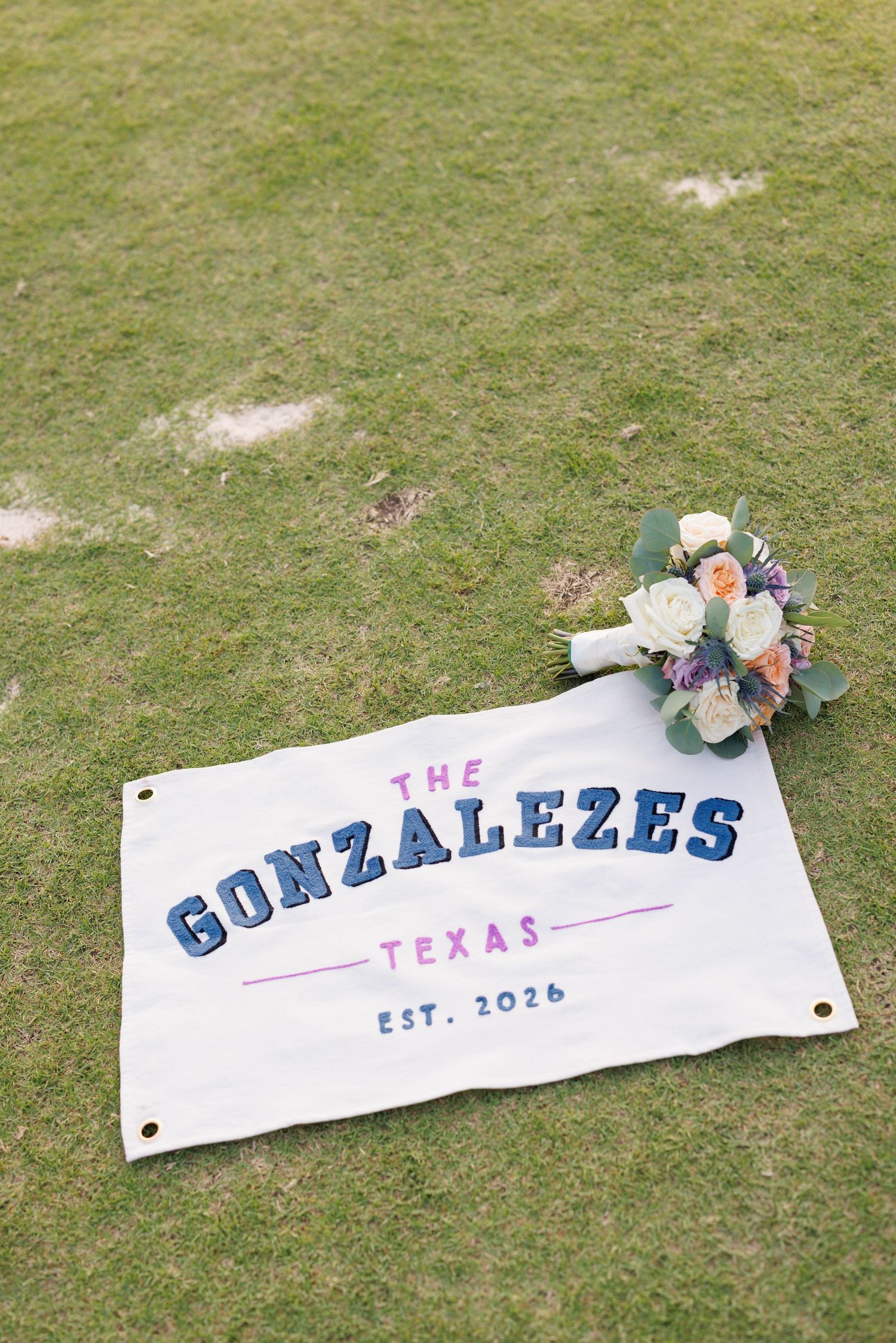 Wedding banner reading The Gonzalezes Texas Est. 2026 laid on grass with a colorful bridal bouquet.