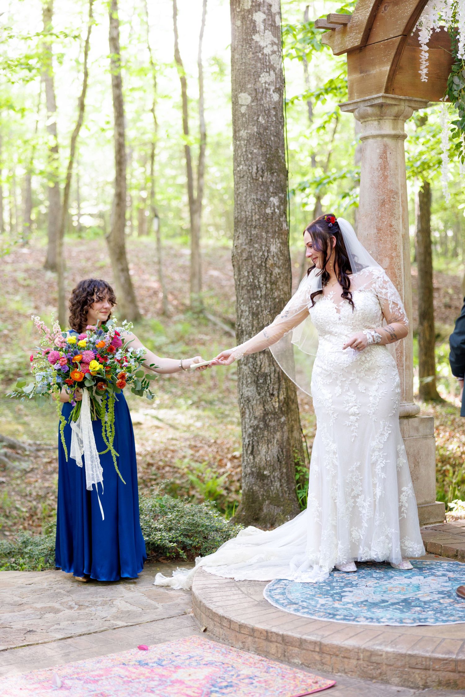Bride in lace wedding gown holds hands with bridesmaid in blue dress at outdoor forest wedding ceremony.