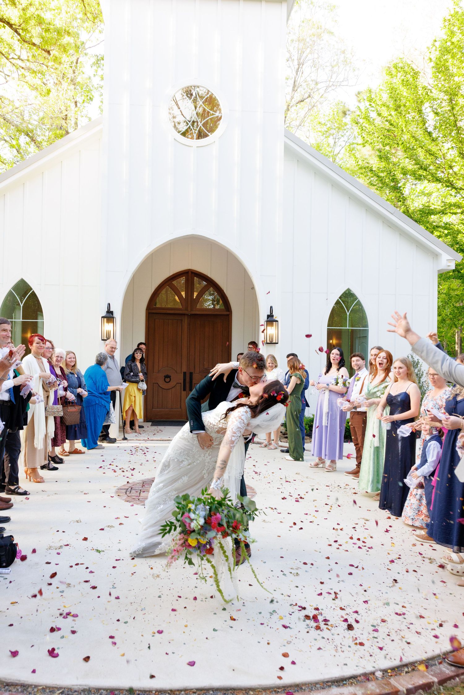 Groom dips bride for romantic kiss outside white chapel as guests toss rose petals in celebration.