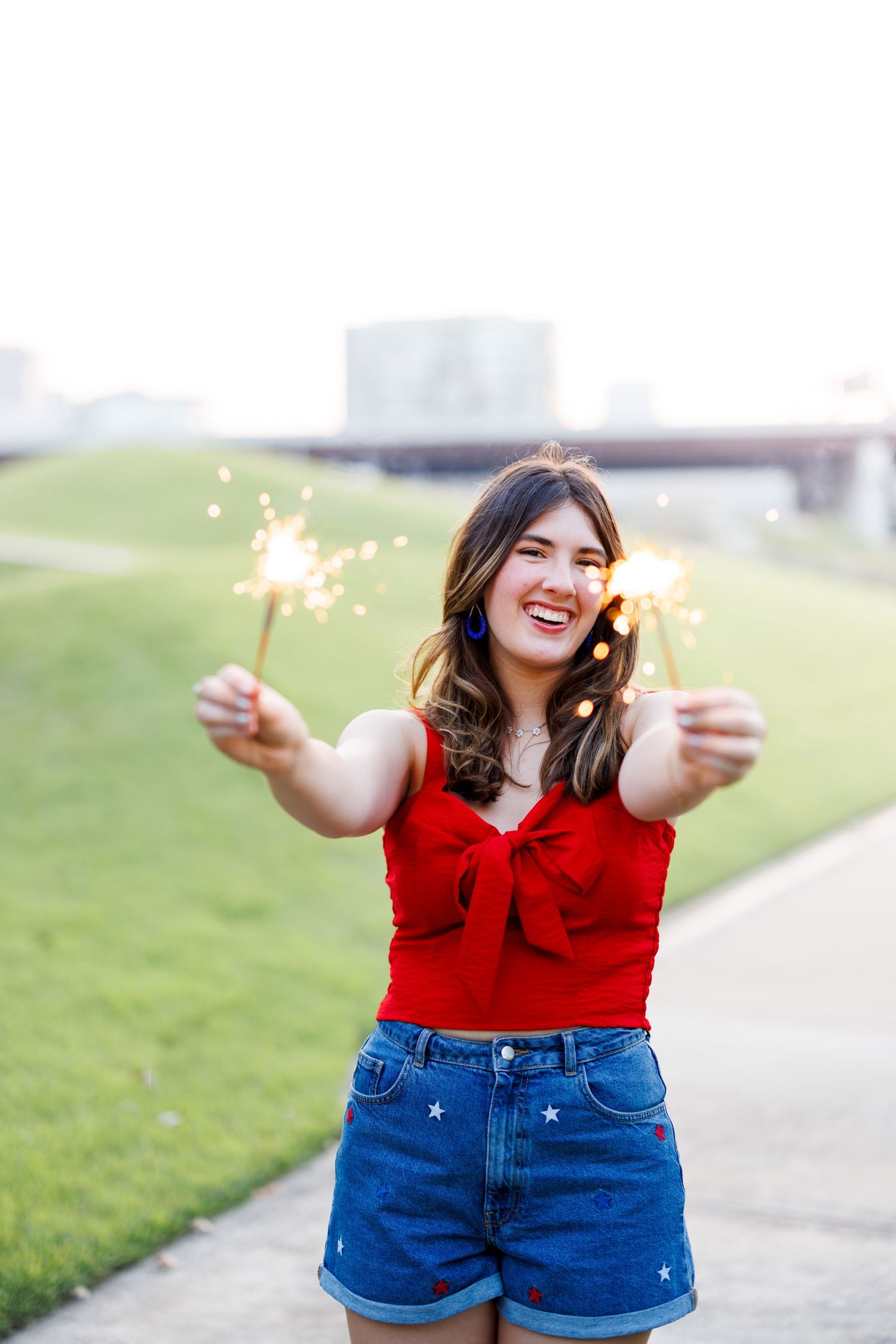 Person in red sleeveless top and denim shorts holding sparklers outdoors during golden hour.