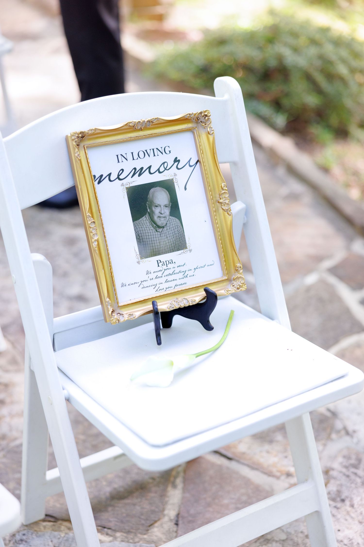 Gold-framed In Loving Memory photo display on white folding chair with white calla lily at outdoor wedding ceremony.