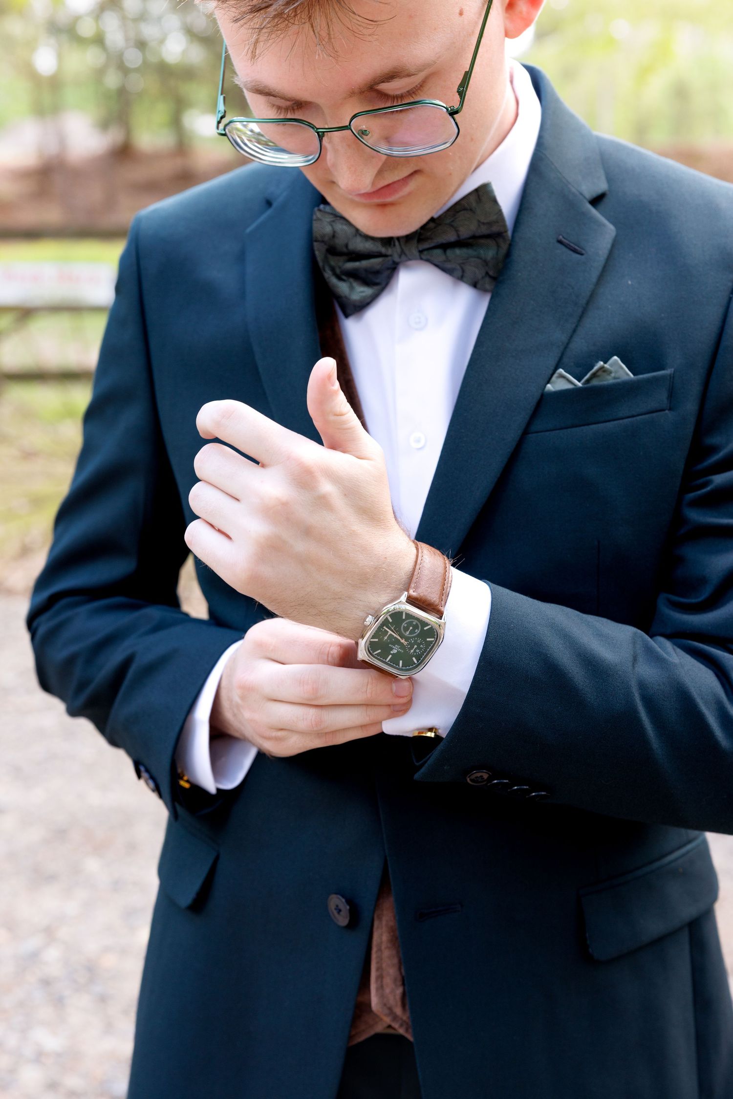 Groom in navy suit adjusting cufflinks with bow tie, glasses, pocket square, and leather watch outdoors.