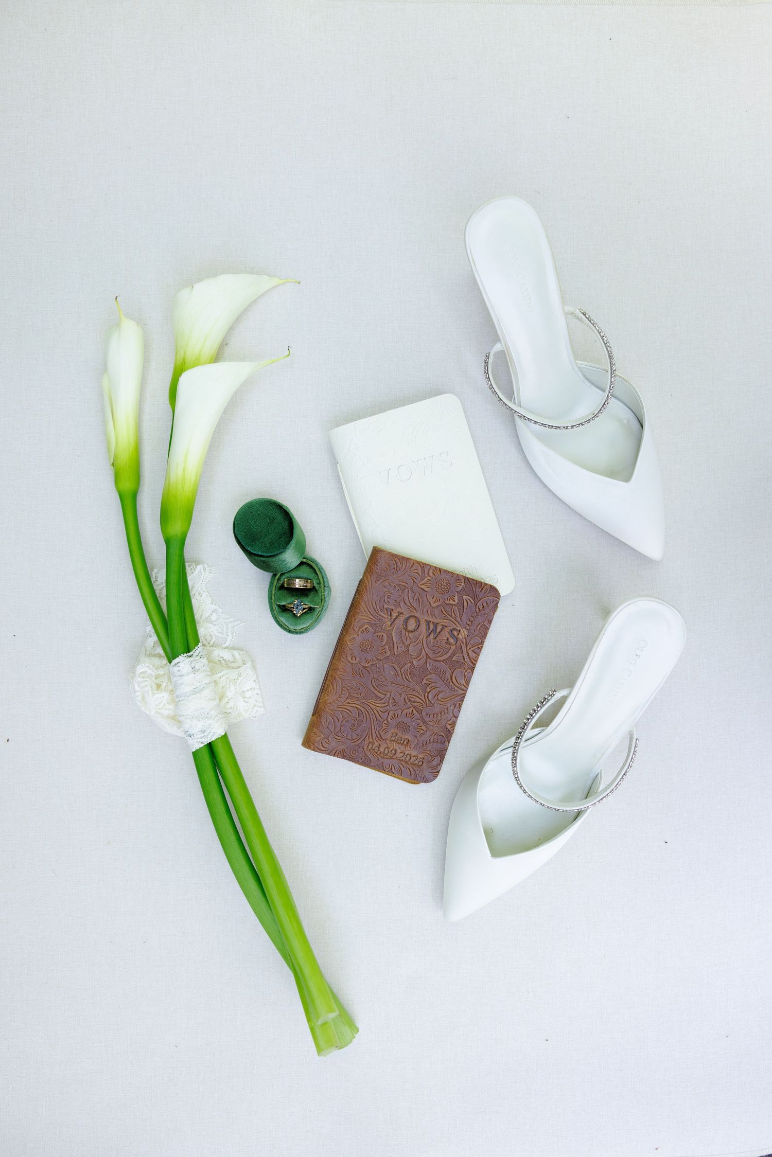 White wedding heels, calla lily bouquet, vow book, and ring box arranged on a white surface for a bridal flat lay.