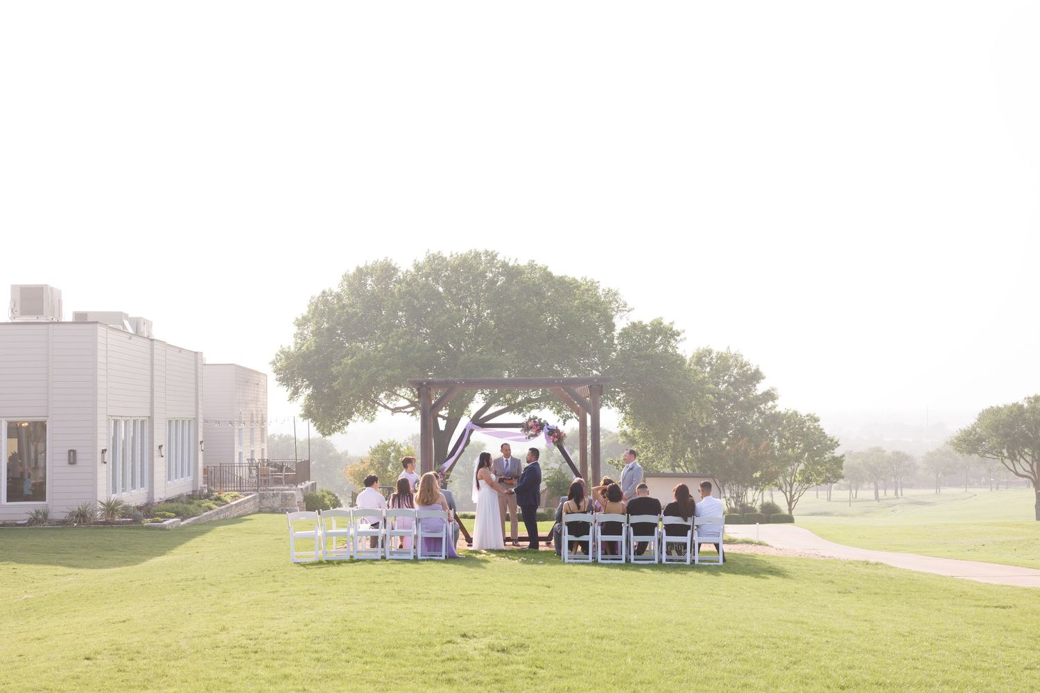 Outdoor wedding ceremony under a wooden pergola on a lush green lawn with guests seated in white chairs.