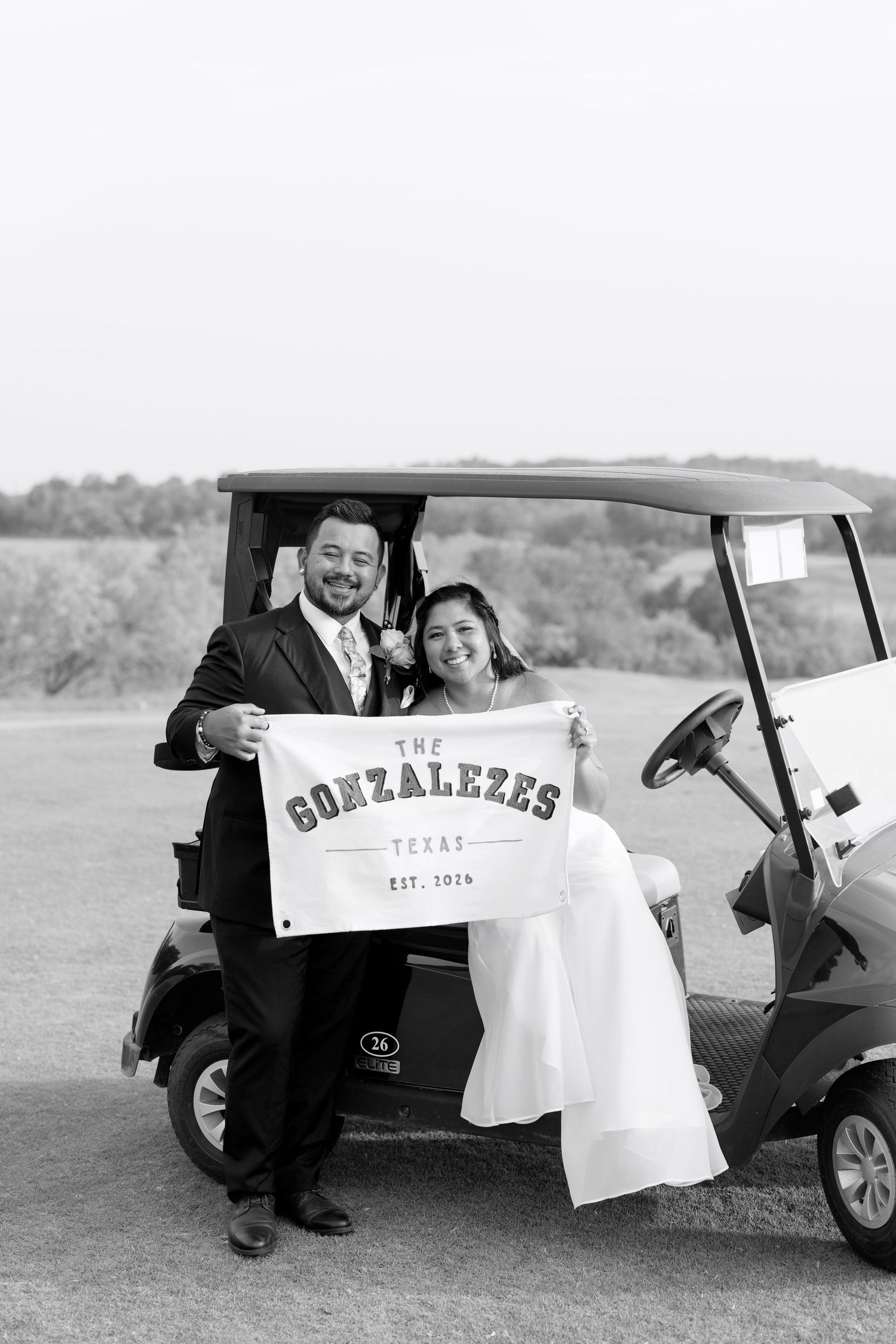 Newlyweds holding a custom Gonzalezes Texas Est. 2024 banner beside a golf cart in black and white.