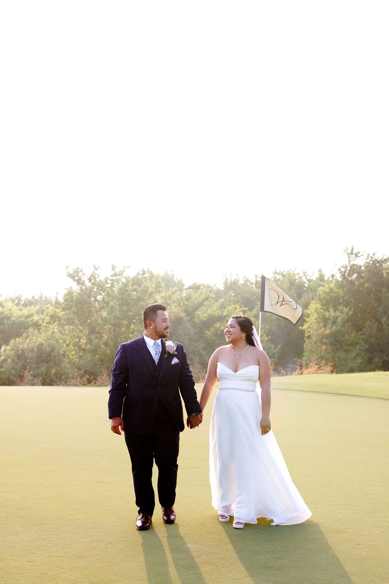 Bride and groom walk hand-in-hand on a golf course at golden hour, smiling at each other in wedding attire.