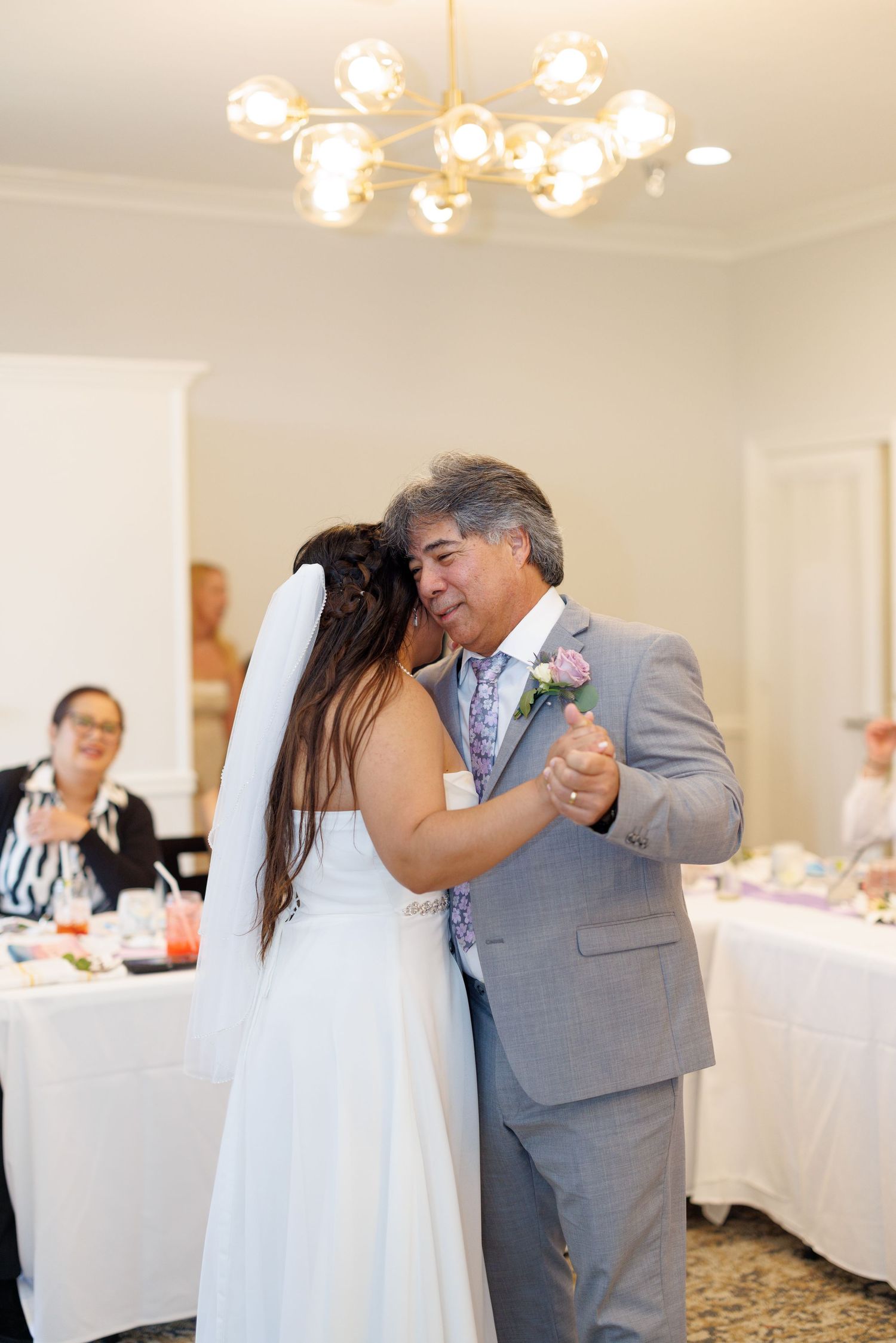Bride and father share an emotional first dance at wedding reception under elegant chandelier lighting.