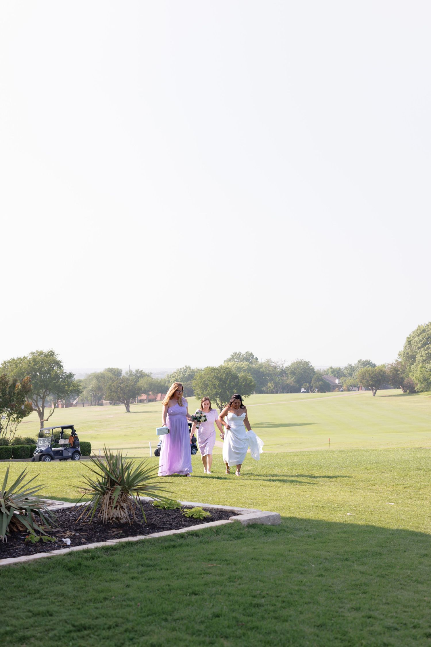 Bridesmaids in lavender dresses walking across a lush green golf course on a sunny wedding day.