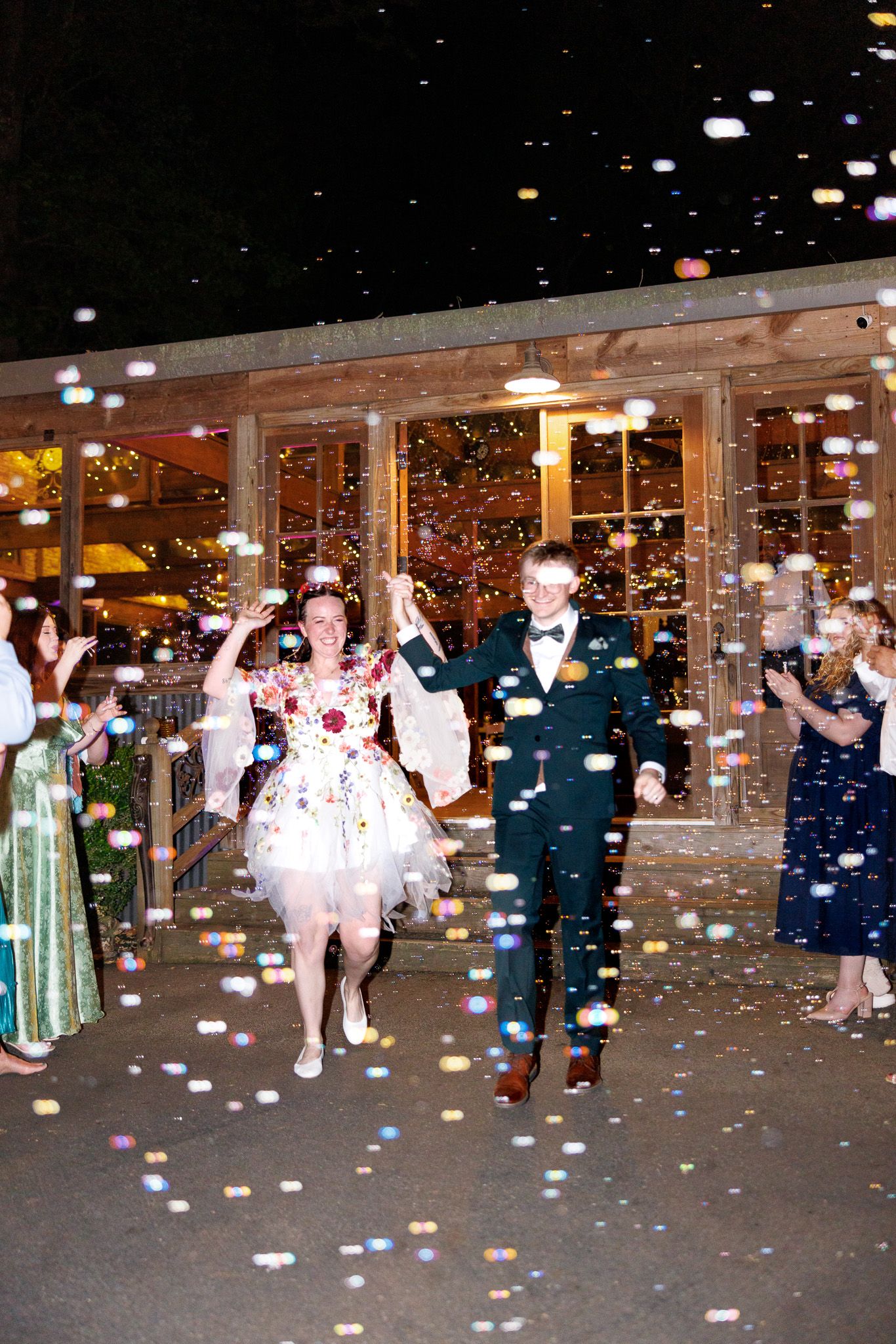 Newlywed couple exits wedding venue through colorful confetti shower as guests celebrate at night.
