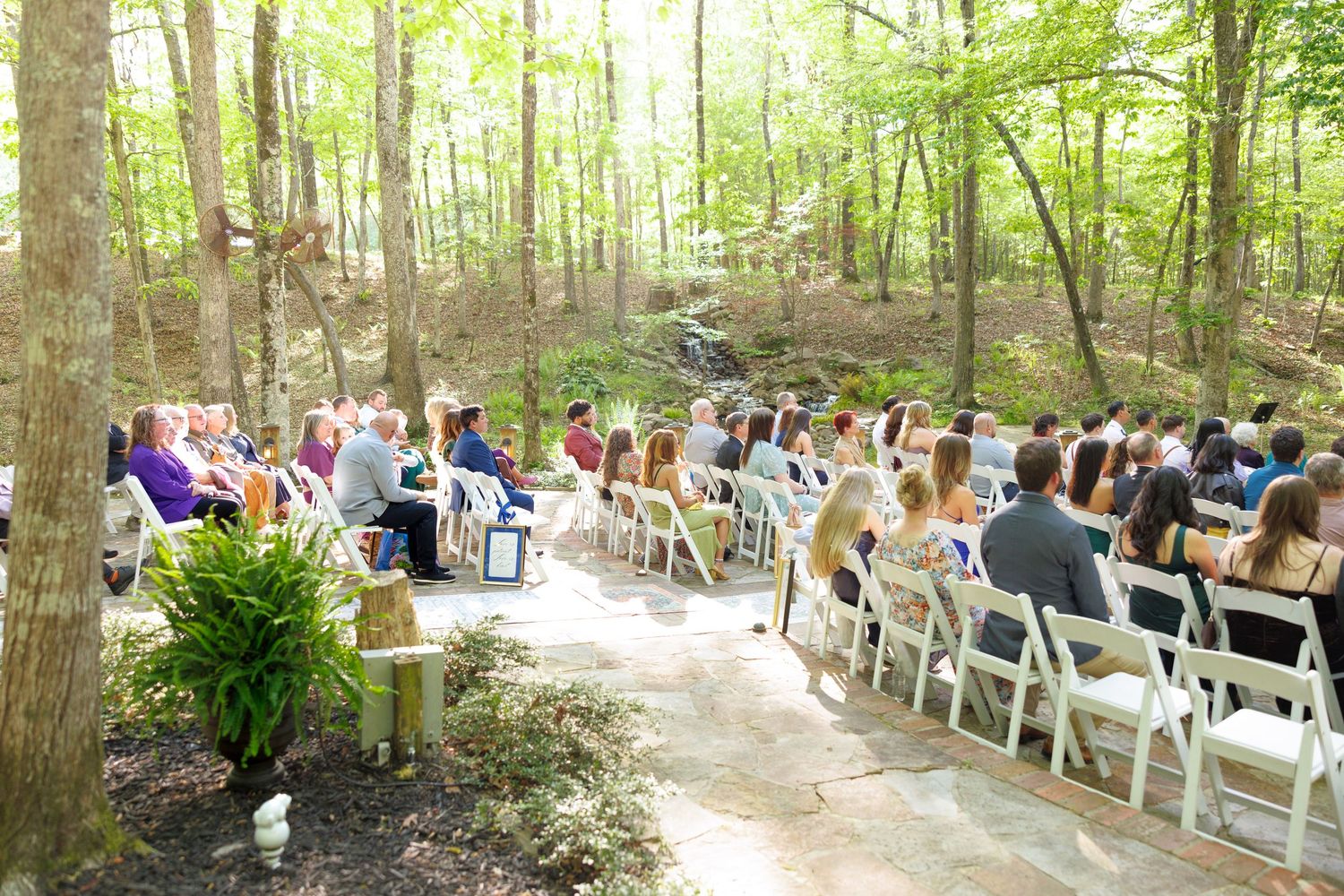 Outdoor woodland wedding ceremony with guests seated in white chairs among tall trees on a sunny day.