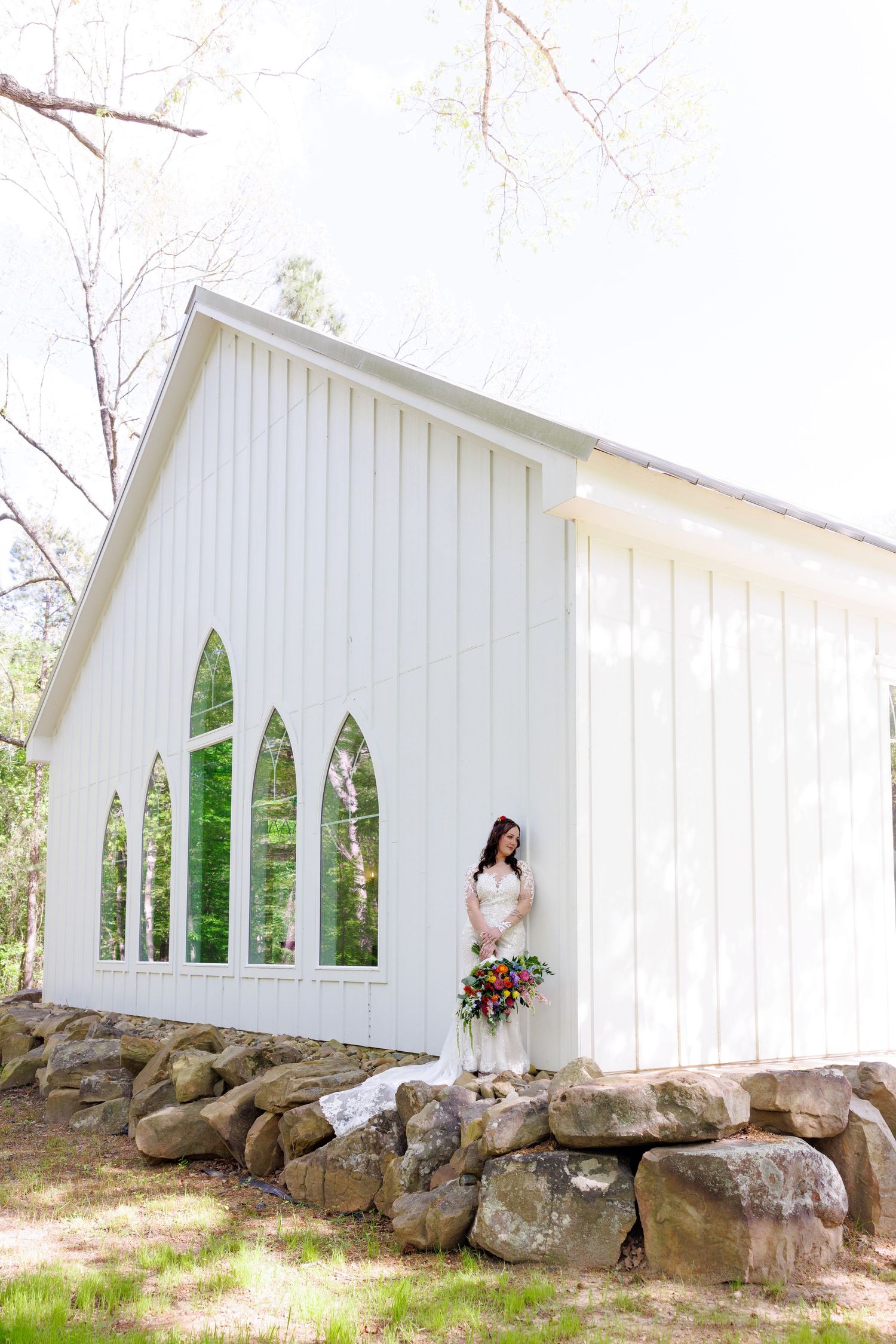 Bride in white lace gown holding colorful bouquet stands beside a white chapel with arched windows amid trees.