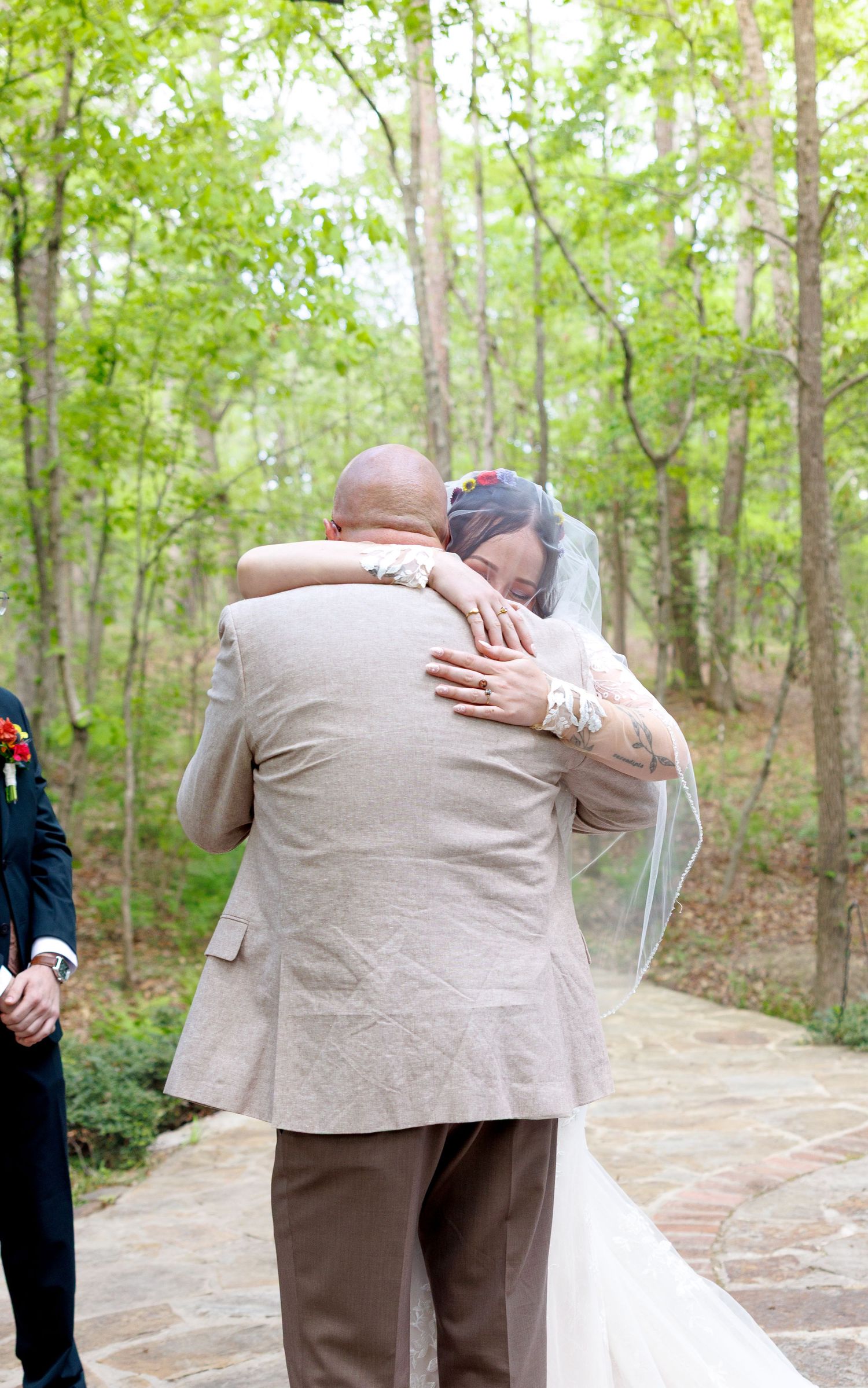 Bride and escort share emotional hug at outdoor wedding ceremony surrounded by lush green forest.