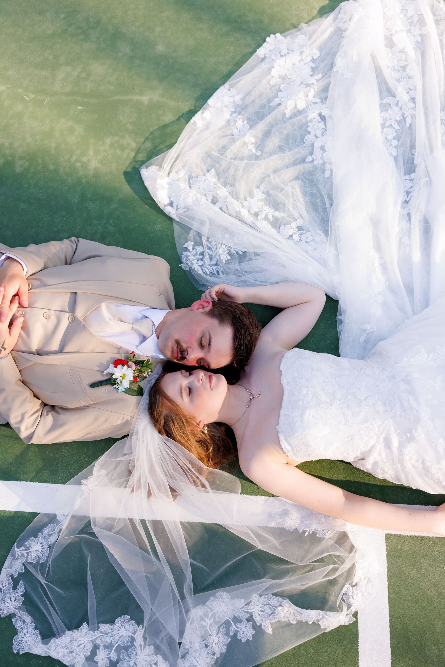 Bride and groom lying on a tennis court, sharing a romantic moment in wedding attire with lace veil and bouquet.
