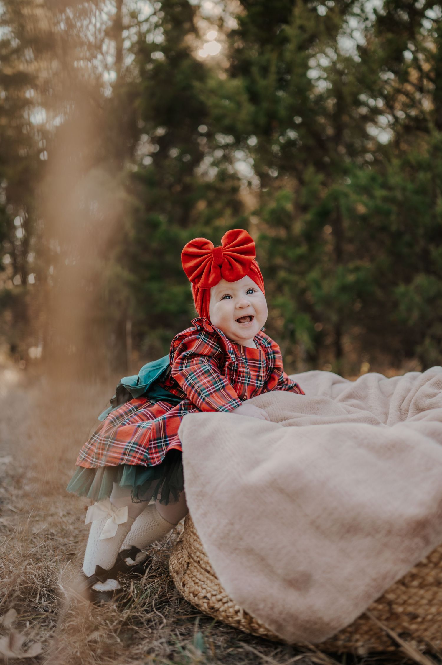 A small child in a plaid dress and red bow headband smiles while playing outdoors near a blanket in the woods.