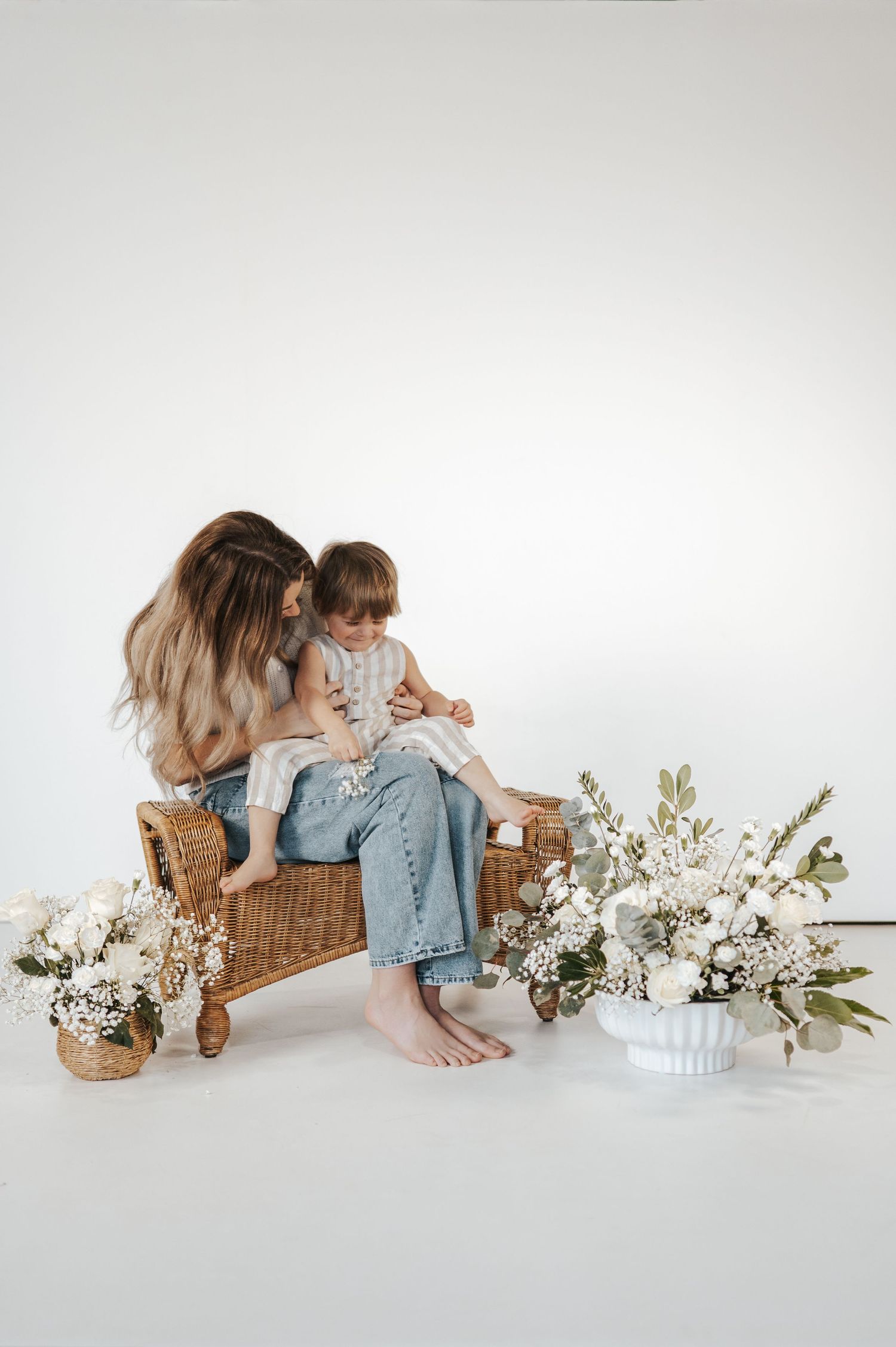 Two people sit together on rattan bench with white floral arrangements in minimalist setting against light backdrop.