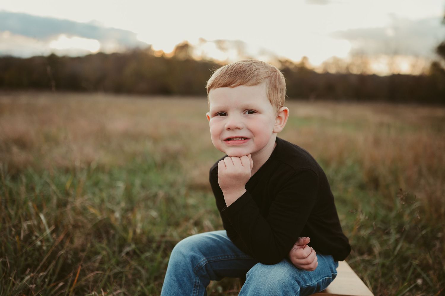 Smiling young boy in black shirt and jeans sitting in a golden field at sunset.