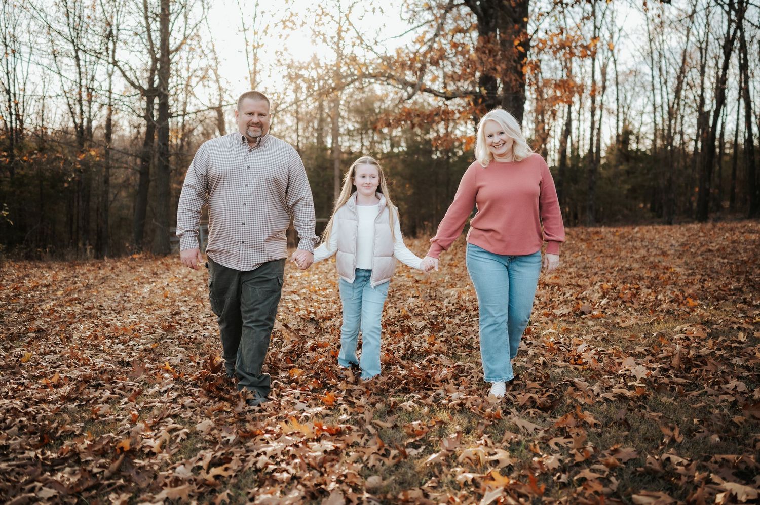 Three people walking hand in hand through fallen autumn leaves in a forest during sunset.