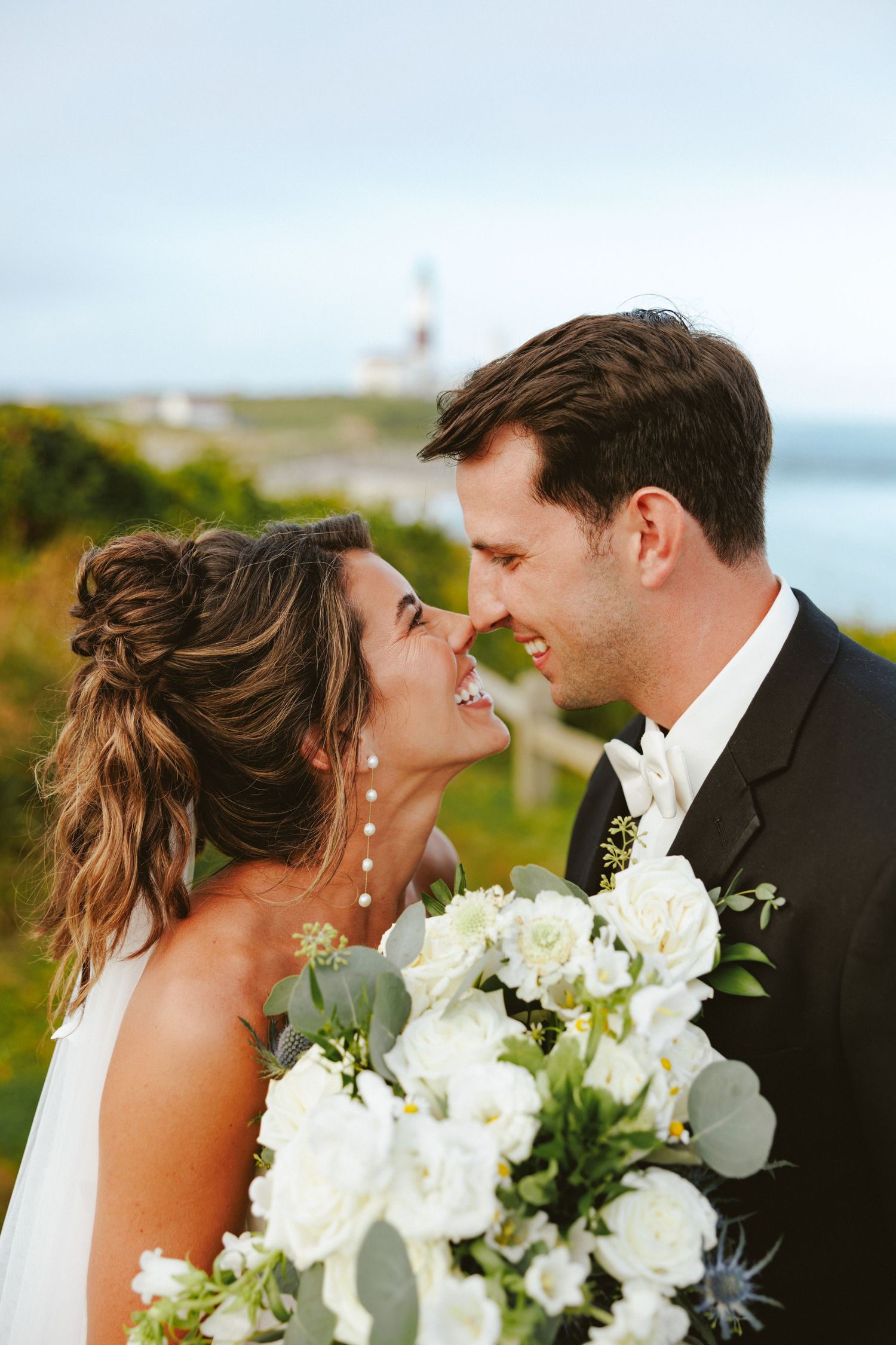 Smiling bride and groom touch noses holding white rose bouquet with coastal lighthouse in background.