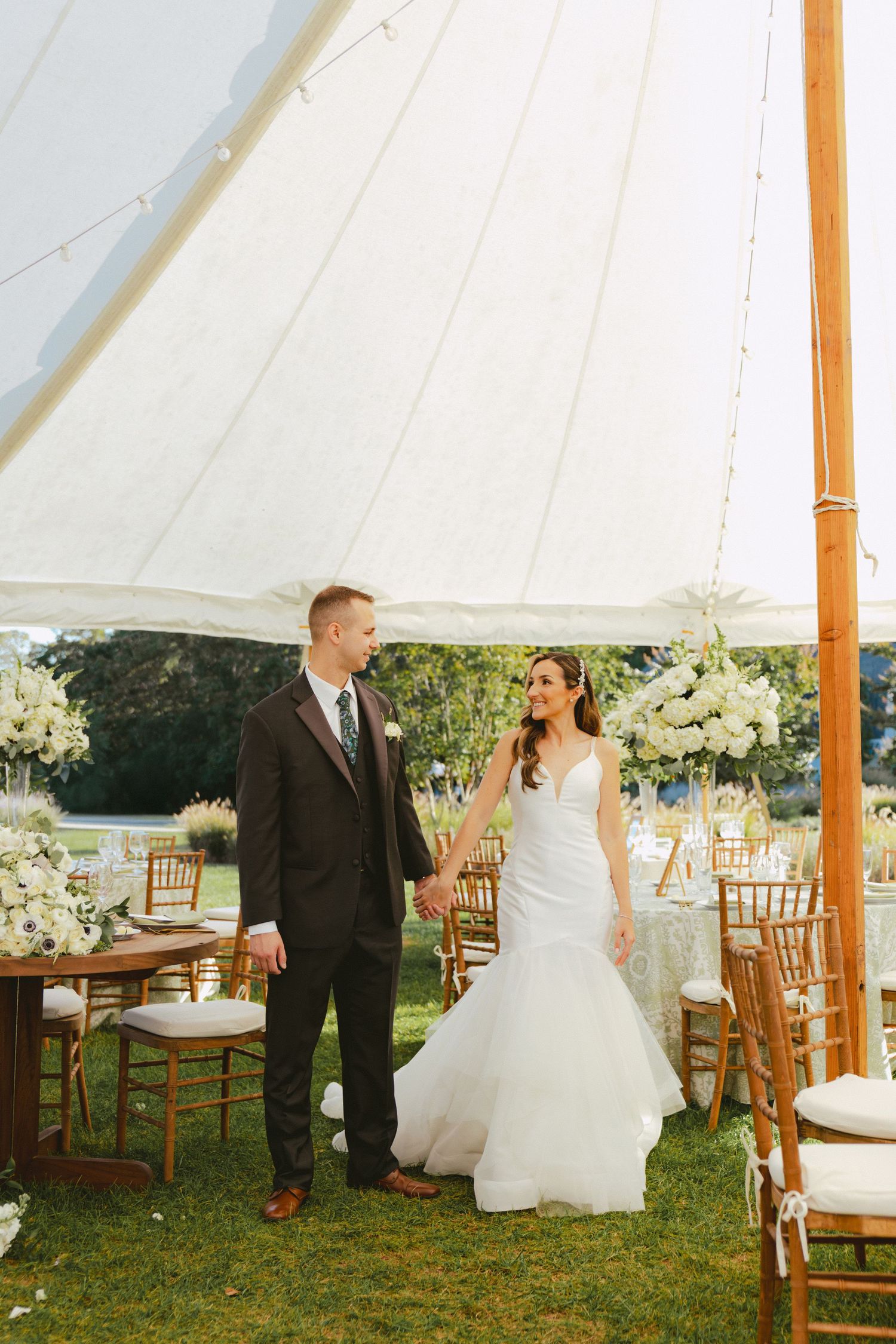 Bride and groom holding hands under white sailcloth tent with floral arrangements at outdoor wedding.