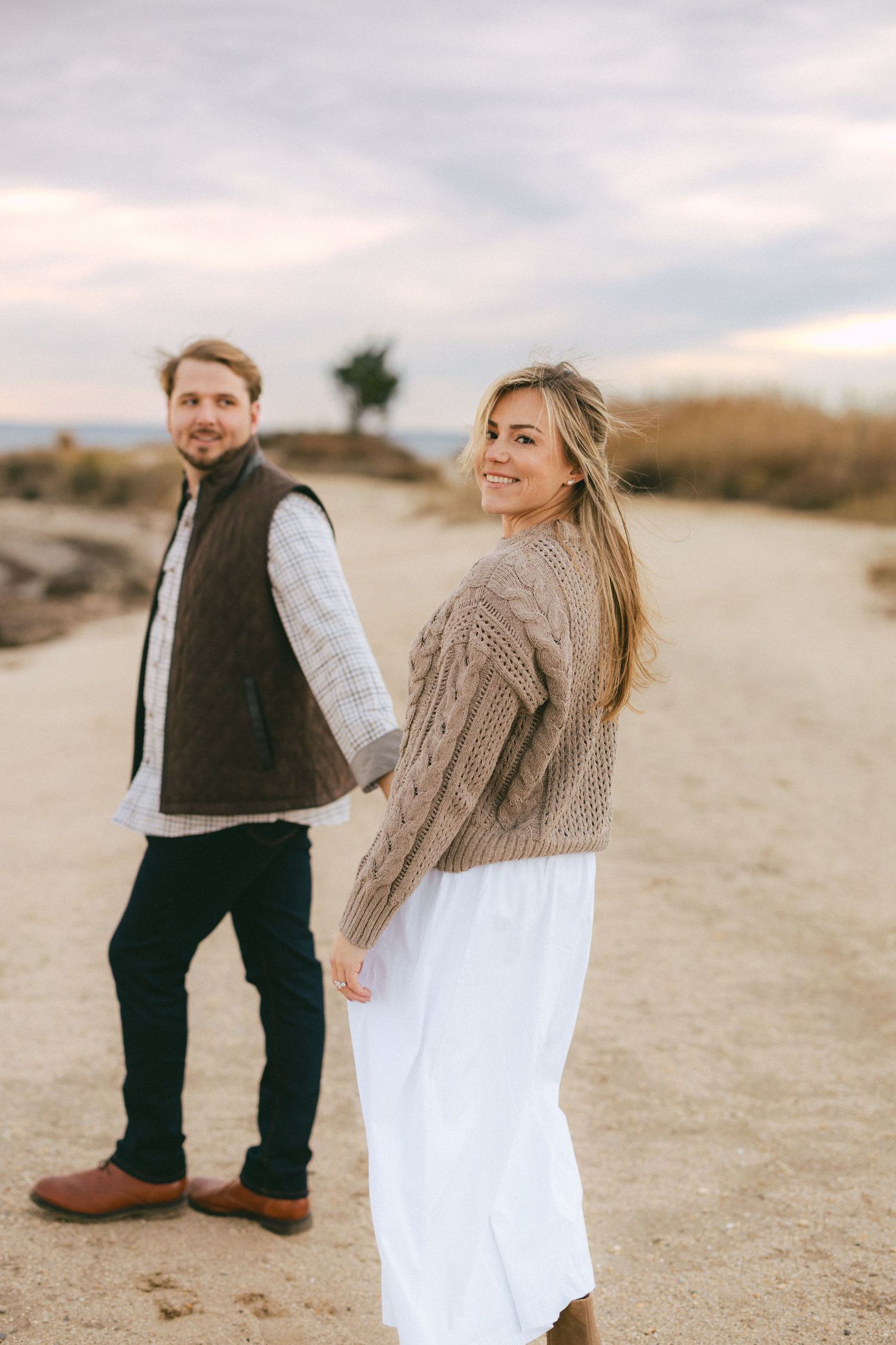 Smiling woman in tan cable knit sweater and white skirt looks back while holding hands with man on sandy path.