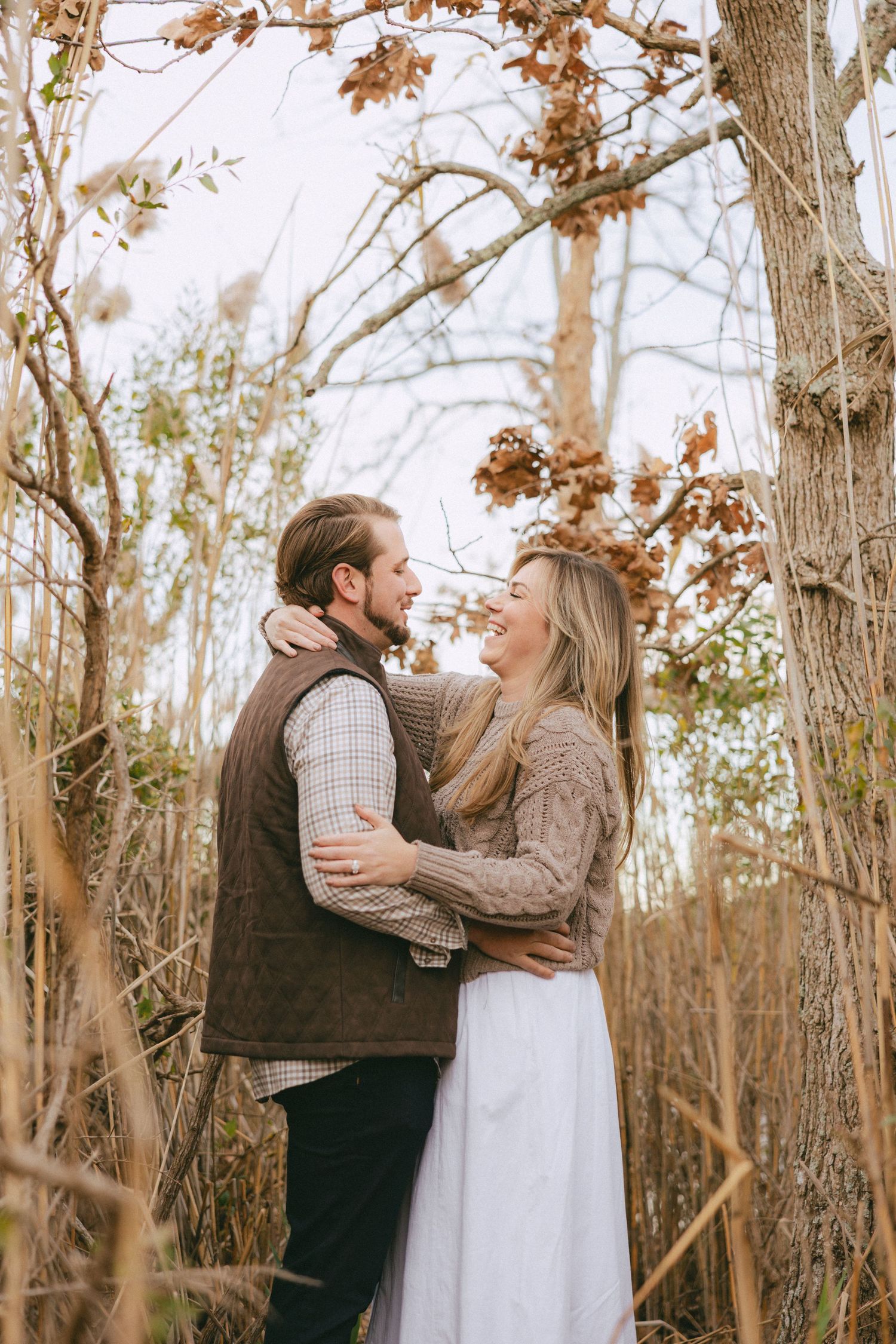 Couple embracing and smiling at each other among bare autumn trees in a natural outdoor setting.
