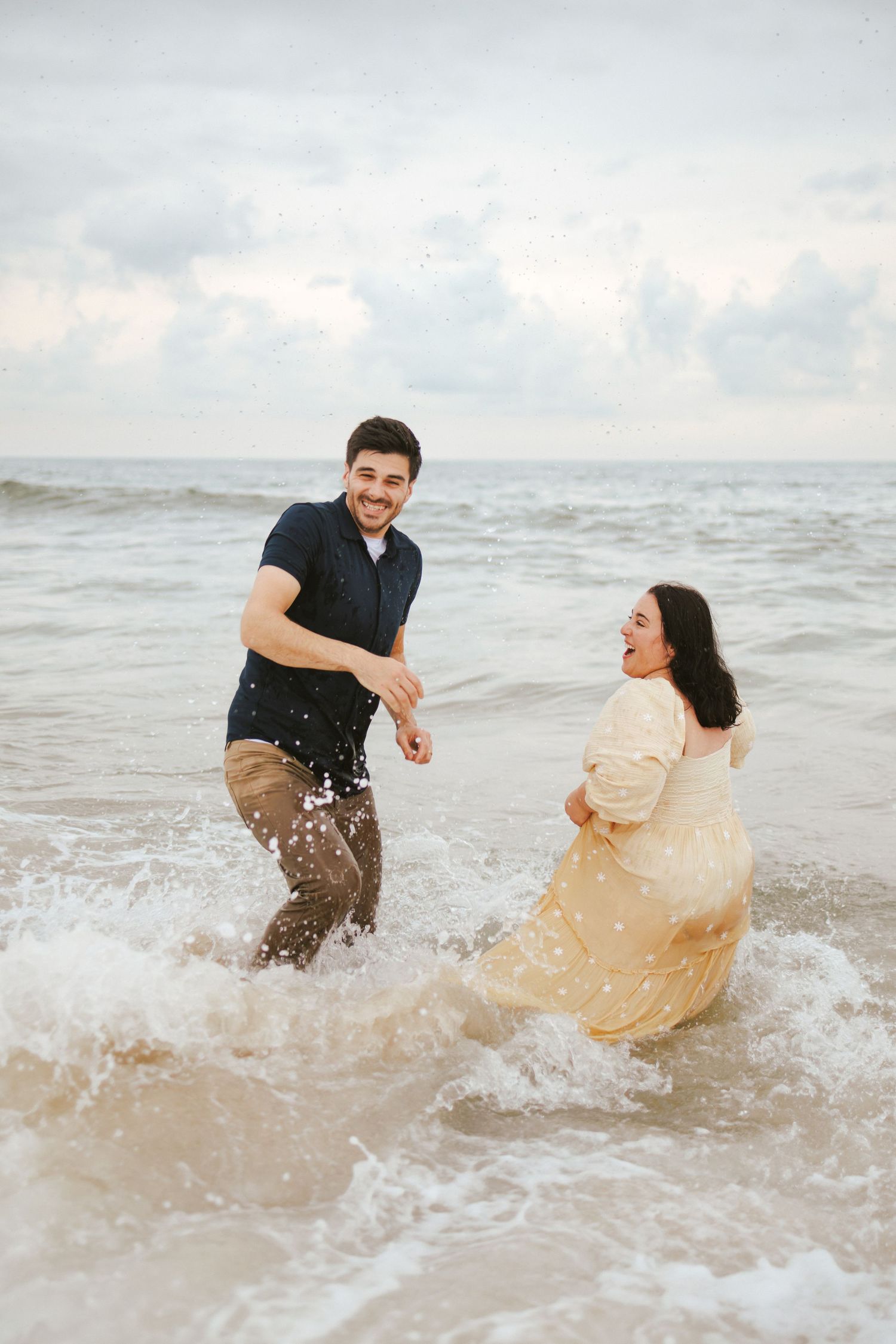 Joyful couple splashes in ocean waves fully clothed during fun beach engagement photo session.