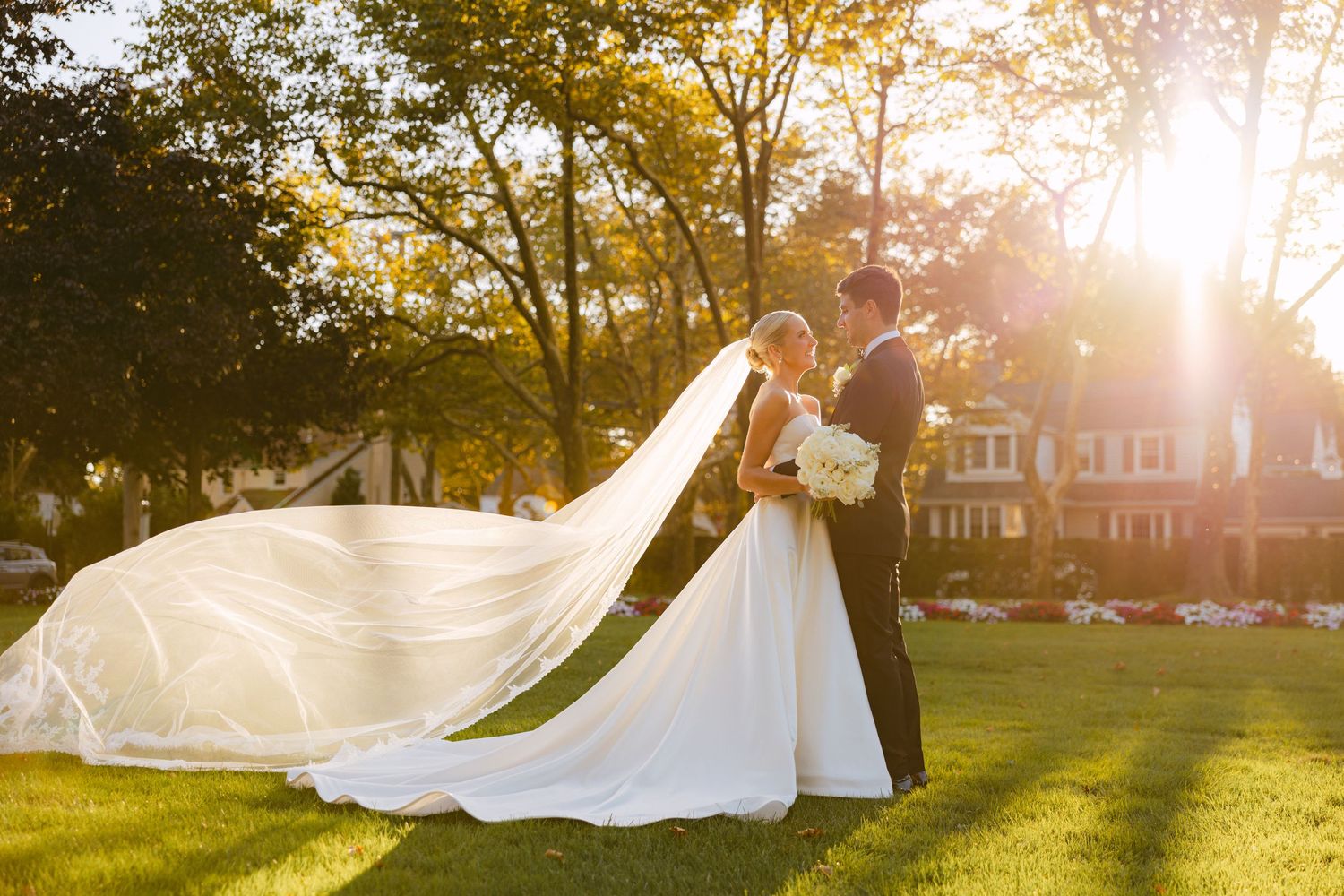 Bride and groom share a romantic sunset moment outdoors, her long veil flowing in the breeze on lush green lawn.