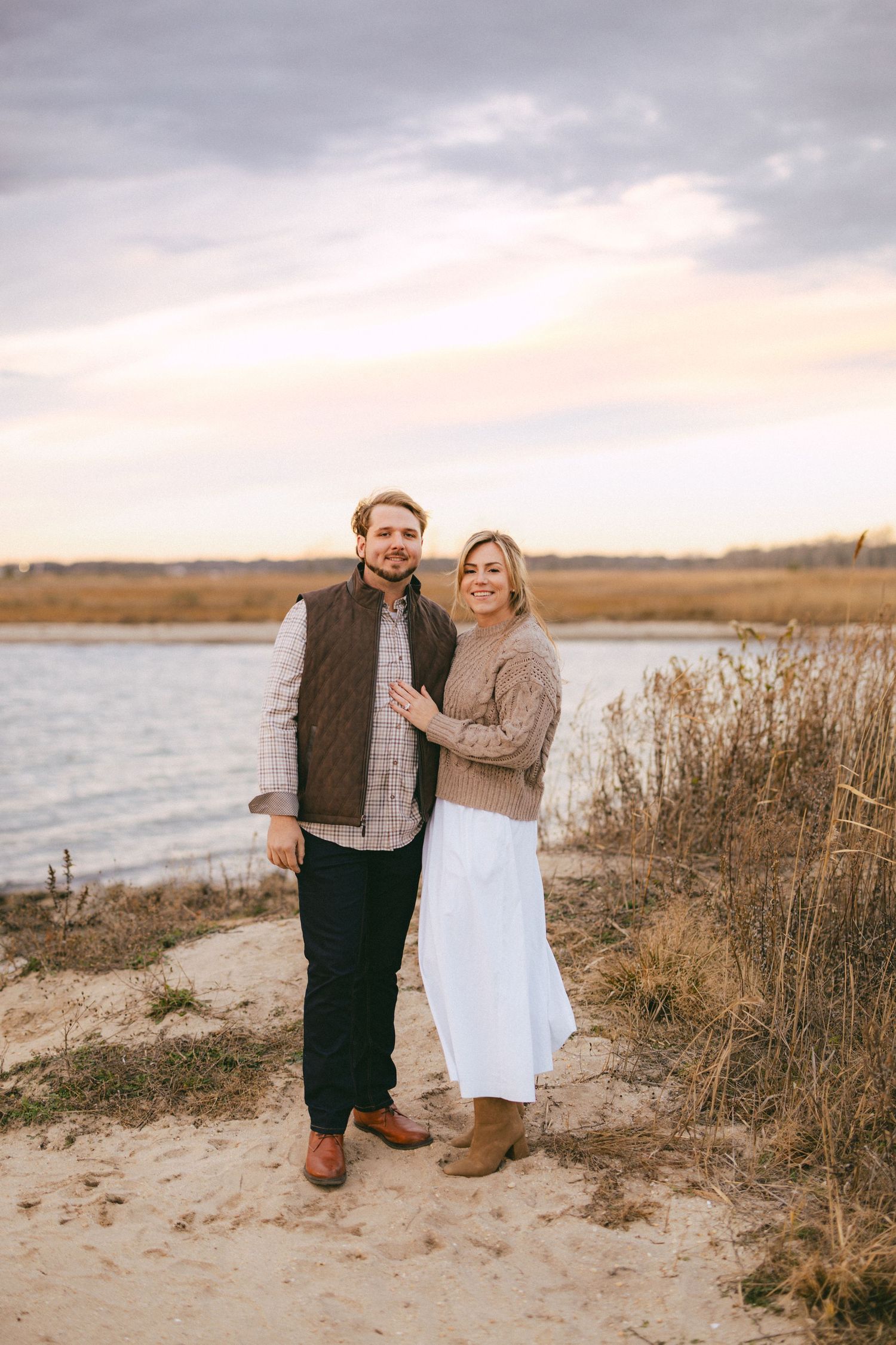 Couple poses together on sandy beach near water at sunset, man in brown vest and woman in white skirt and tan sweater.