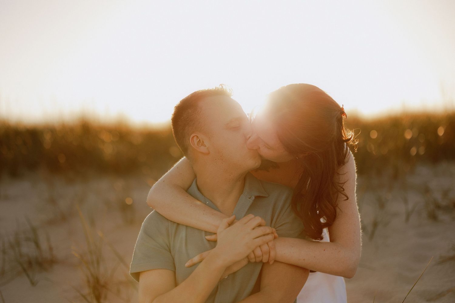 Couple sharing a golden sunset kiss on sandy beach dunes, bathed in warm backlit sunlight.