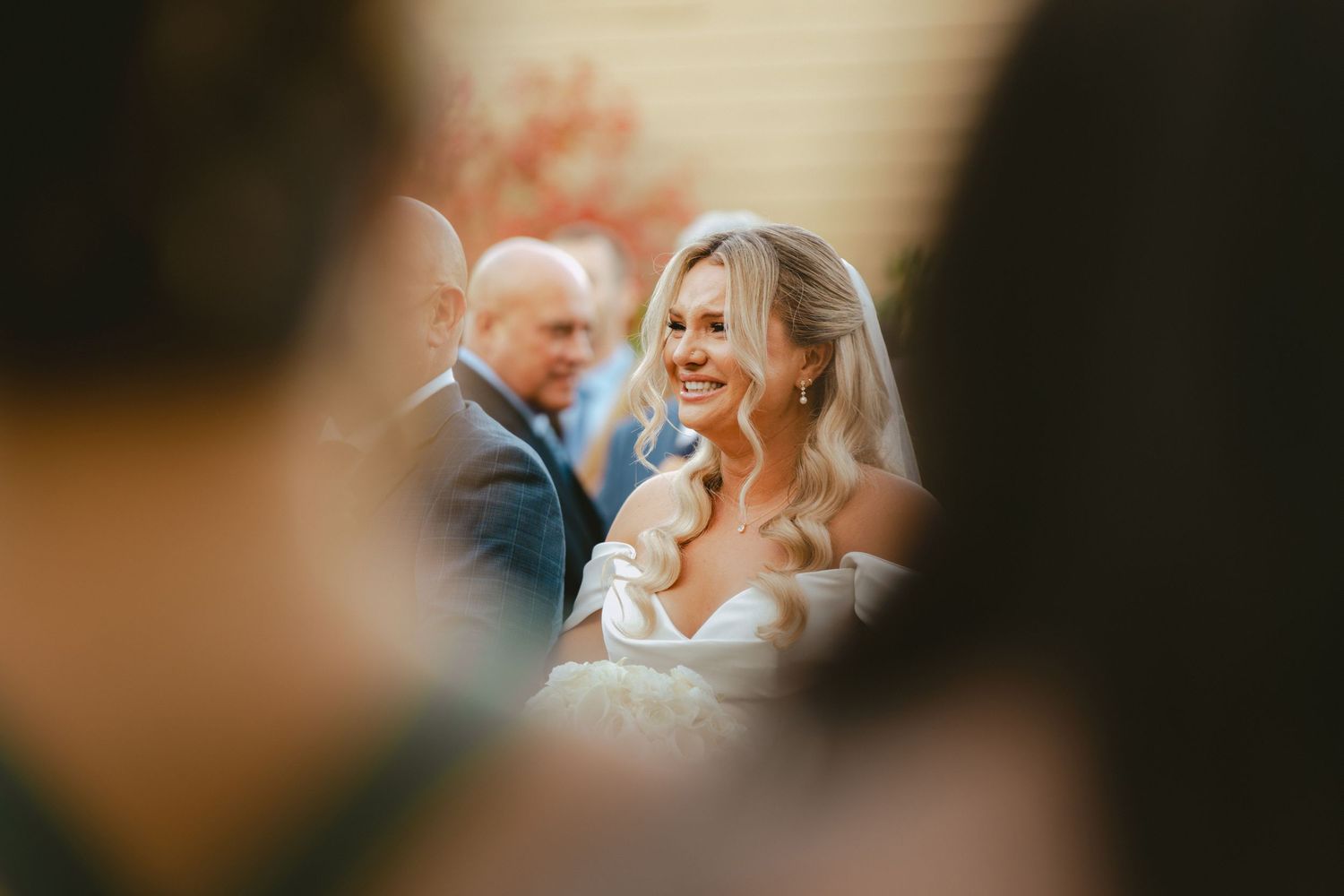 Radiant bride smiling joyfully during wedding ceremony, captured through a soft foreground frame.