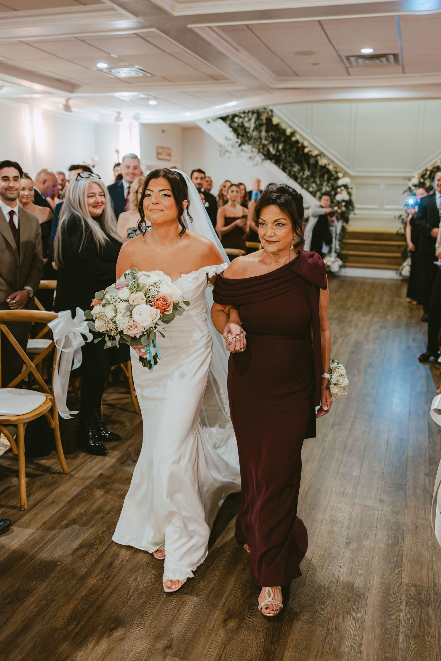 Bride in off-shoulder gown walks down aisle with mother in burgundy dress at indoor wedding.